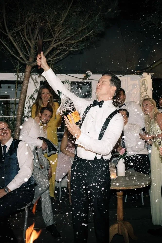 Hochzeitsparty bei Nacht mit einer Gruppe von Menschen, die feiern, ein Mann in Fliege schwingt eine Axt, um Konfetti zu werfen, während andere in festlicher Kleidung lachen und zusehen.