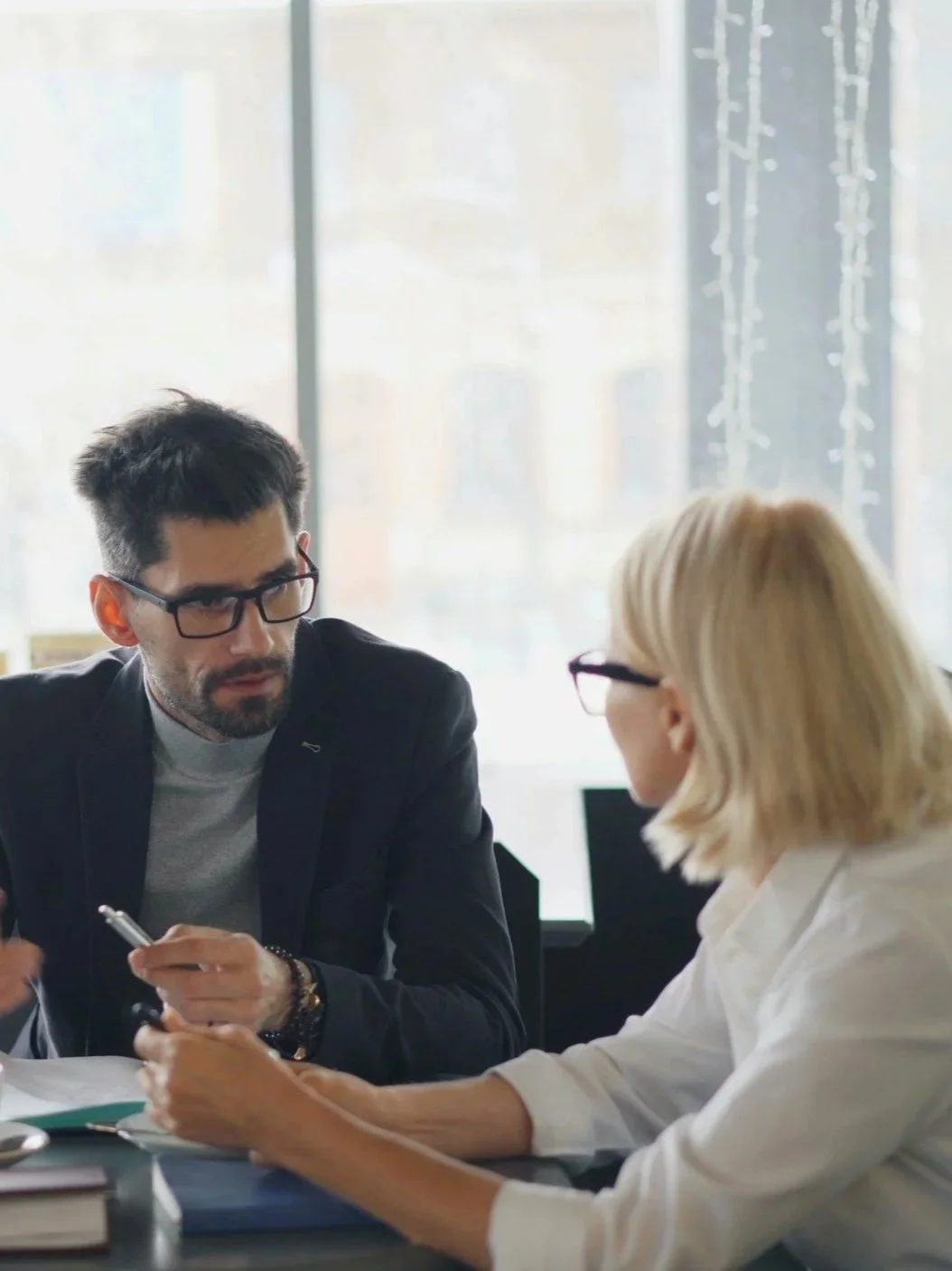 A man with dark hair and glasses sitting across from a blonde woman with glasses at a table in an office or conference room, engaged in a discussion.