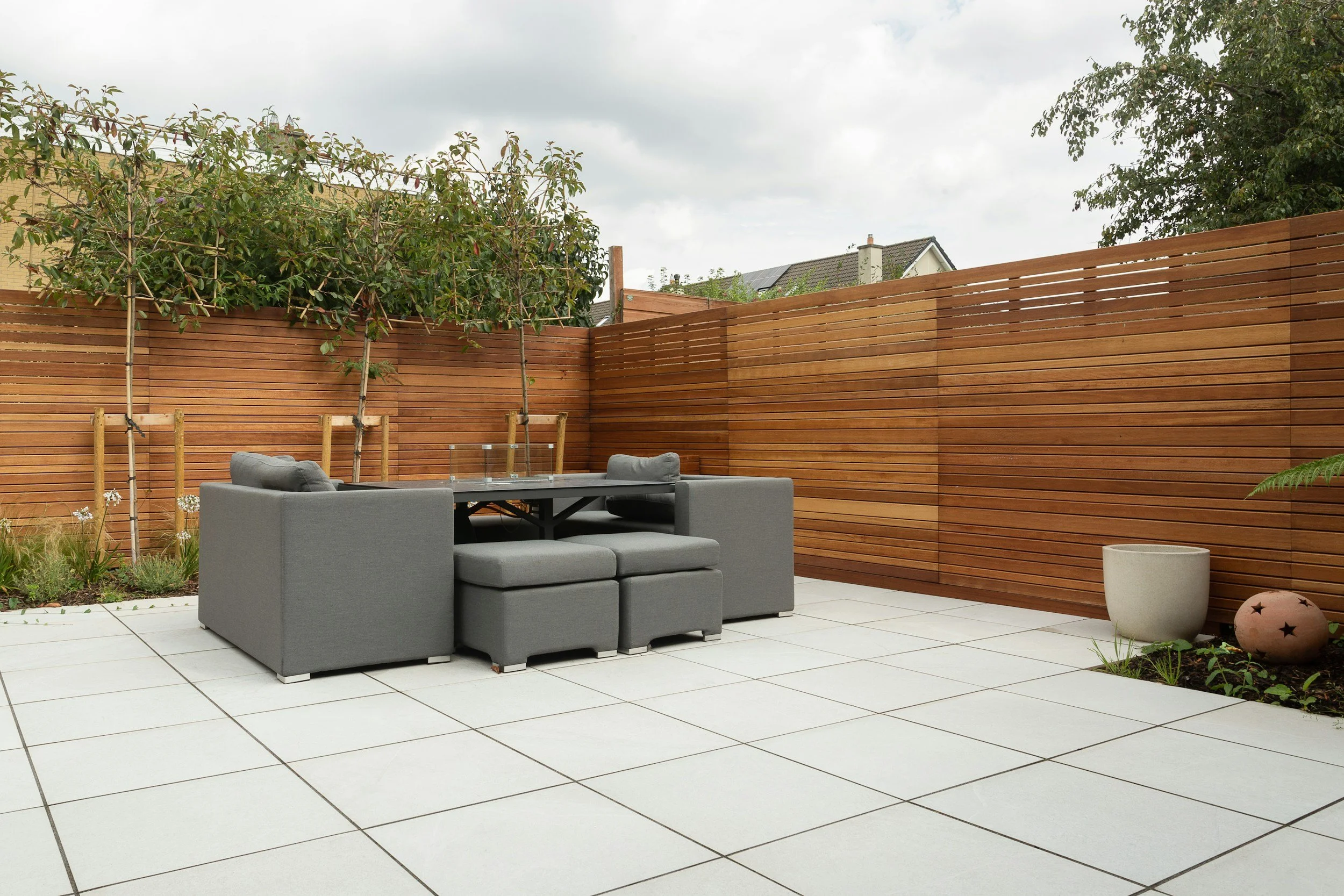A modern backyard patio with a black picnic table, gray outdoor sofa, and potted plants, enclosed by a tall wooden fence and trees with a cloudy sky overhead.
