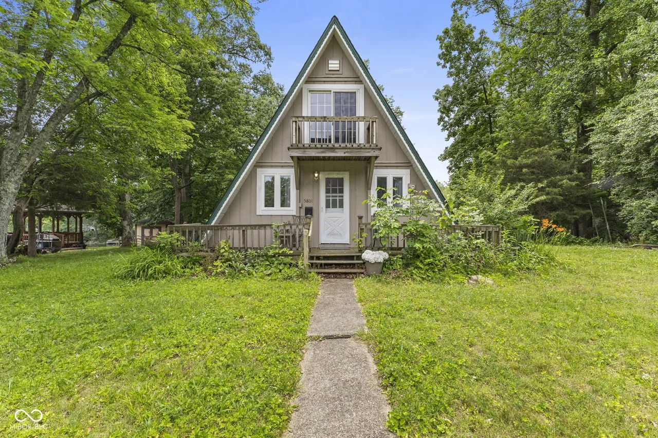 Front view of a tan A-frame home with an upper balcony and wooden deck, surrounded by mature trees.