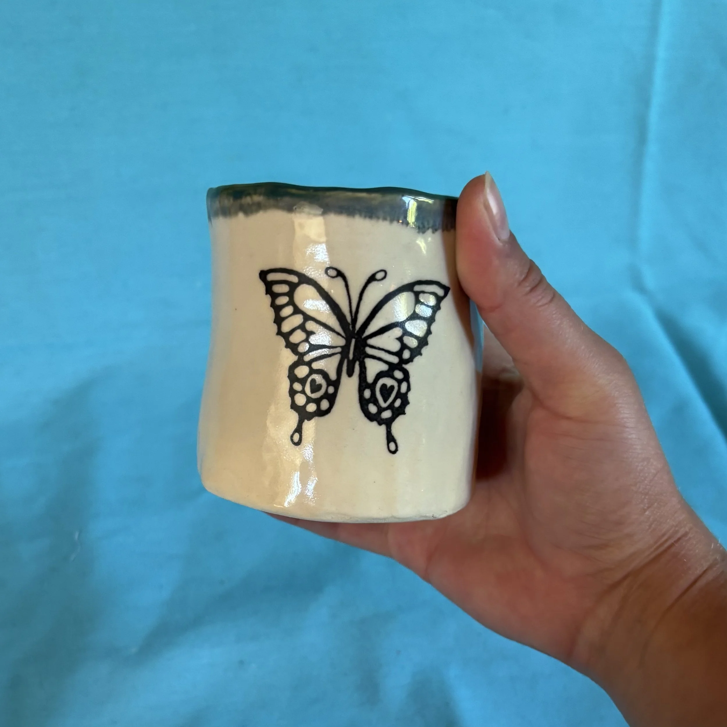 A hand holding a white ceramic mug with a black butterfly design against a plain blue background.