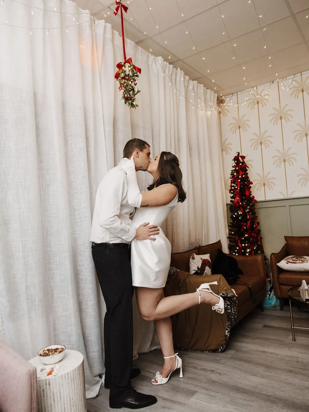 A couple dressed in formal attire sharing a kiss indoors decorated for Christmas with a small Christmas tree, a Christmas pillow, and festive decorations.