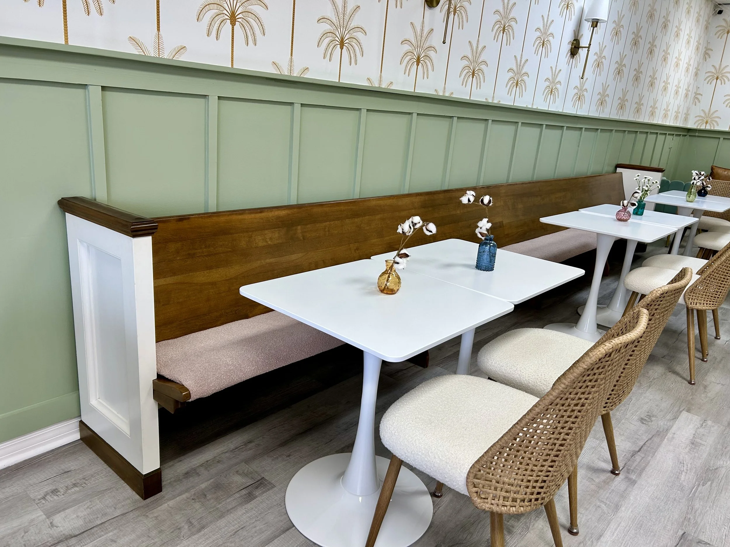 seating area with church pew with dusty rose pink seat, refinished wood, comfy cream chairs, colored vases with small flowers, mint board and batten wall with tropical wall paper, cream with gold palm trees.