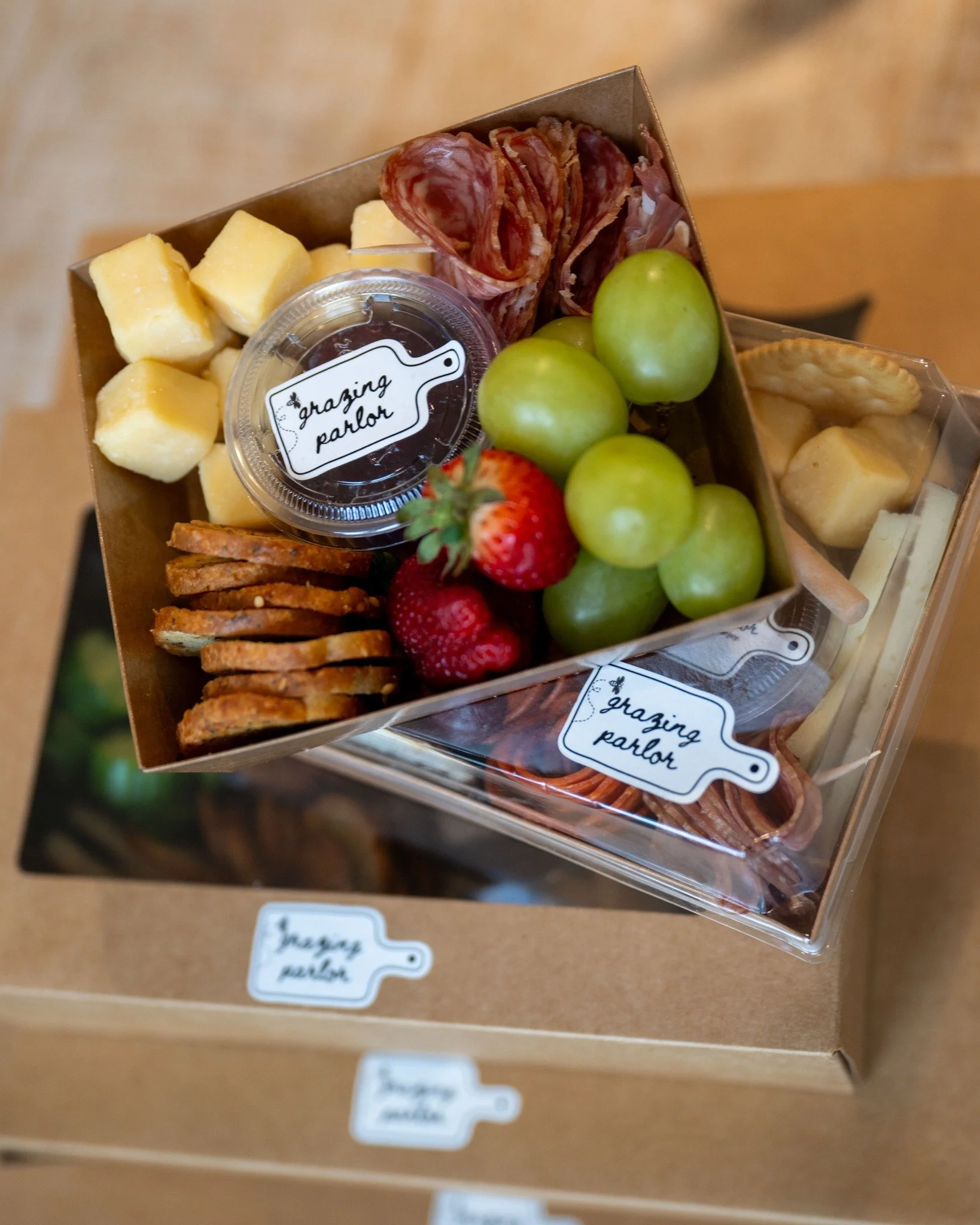 A gift box with assorted fruits, cheese cubes, sliced cured meat, crackers, and a small container of dipping sauce, all labeled 'grazing parlor'.