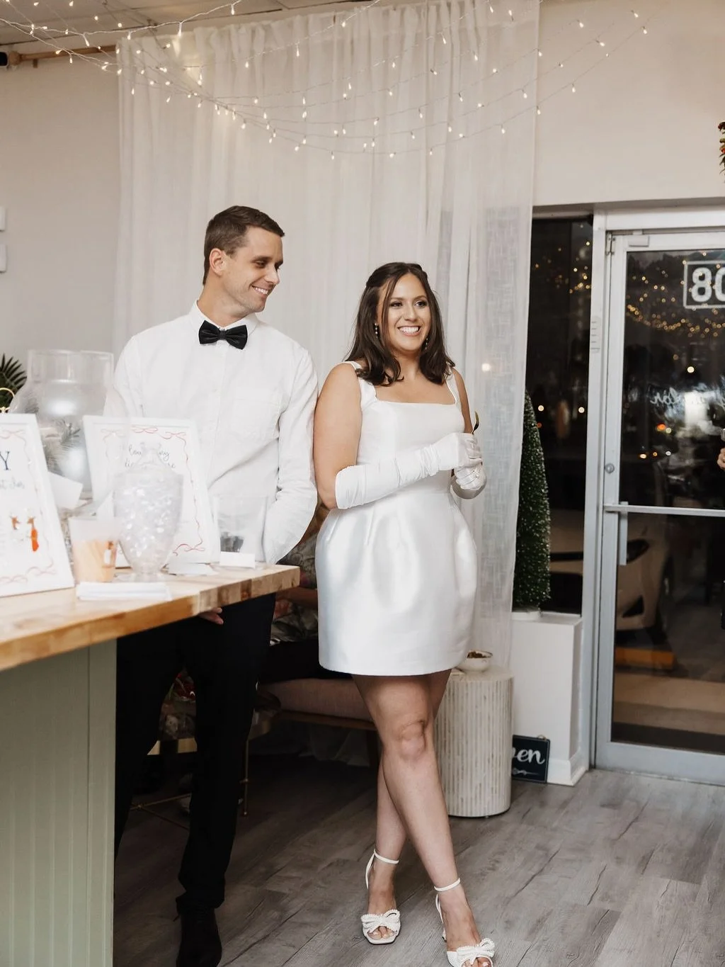 A man and woman dressed in formal attire standing inside a decorated venue, possibly for a special event. The man wears a white shirt with a black bow tie, and the woman wears a white dress with matching white gloves and high heels. They are smiling and standing near a table with decorative items, and string lights hang from the ceiling.