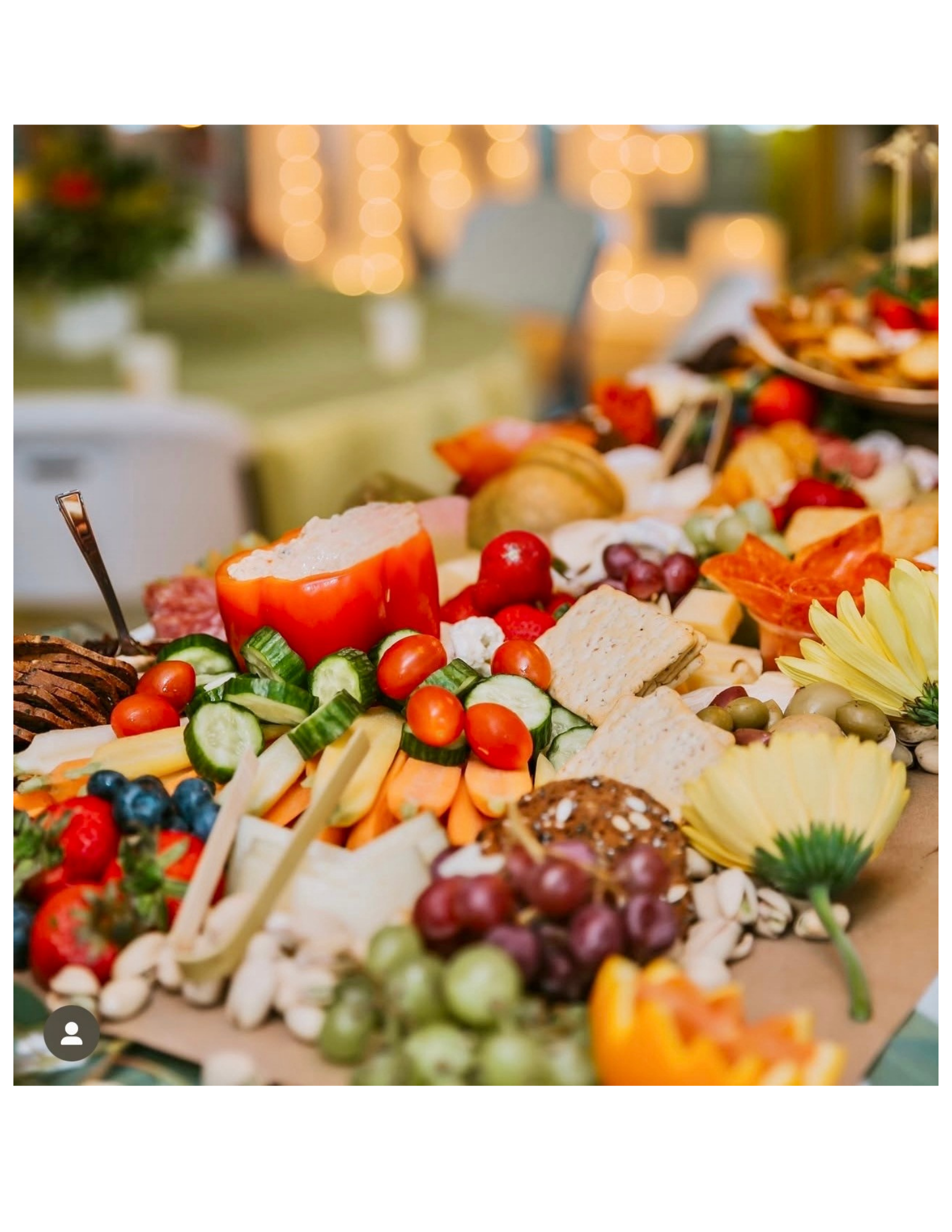 Assorted cheese, fresh vegetables, grapes, strawberries, and crackers on a buffet table with blurred holiday lights in the background.