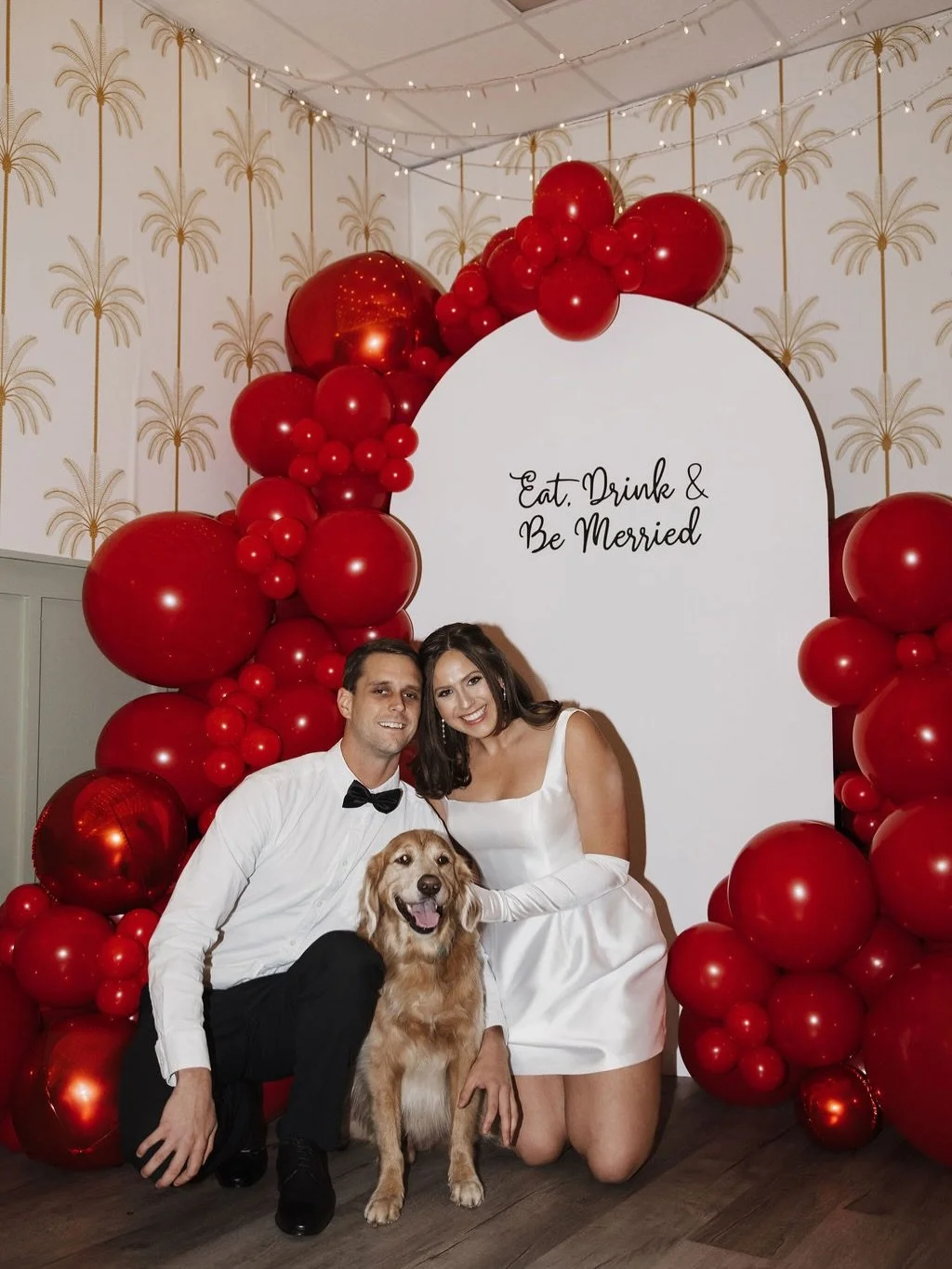 A couple in wedding attire and a dog are posing in front of a decorated backdrop that says "Eat, Drink & Be Married." The backdrop is adorned with large red balloons, and the couple is smiling happily inside a wedding celebration.