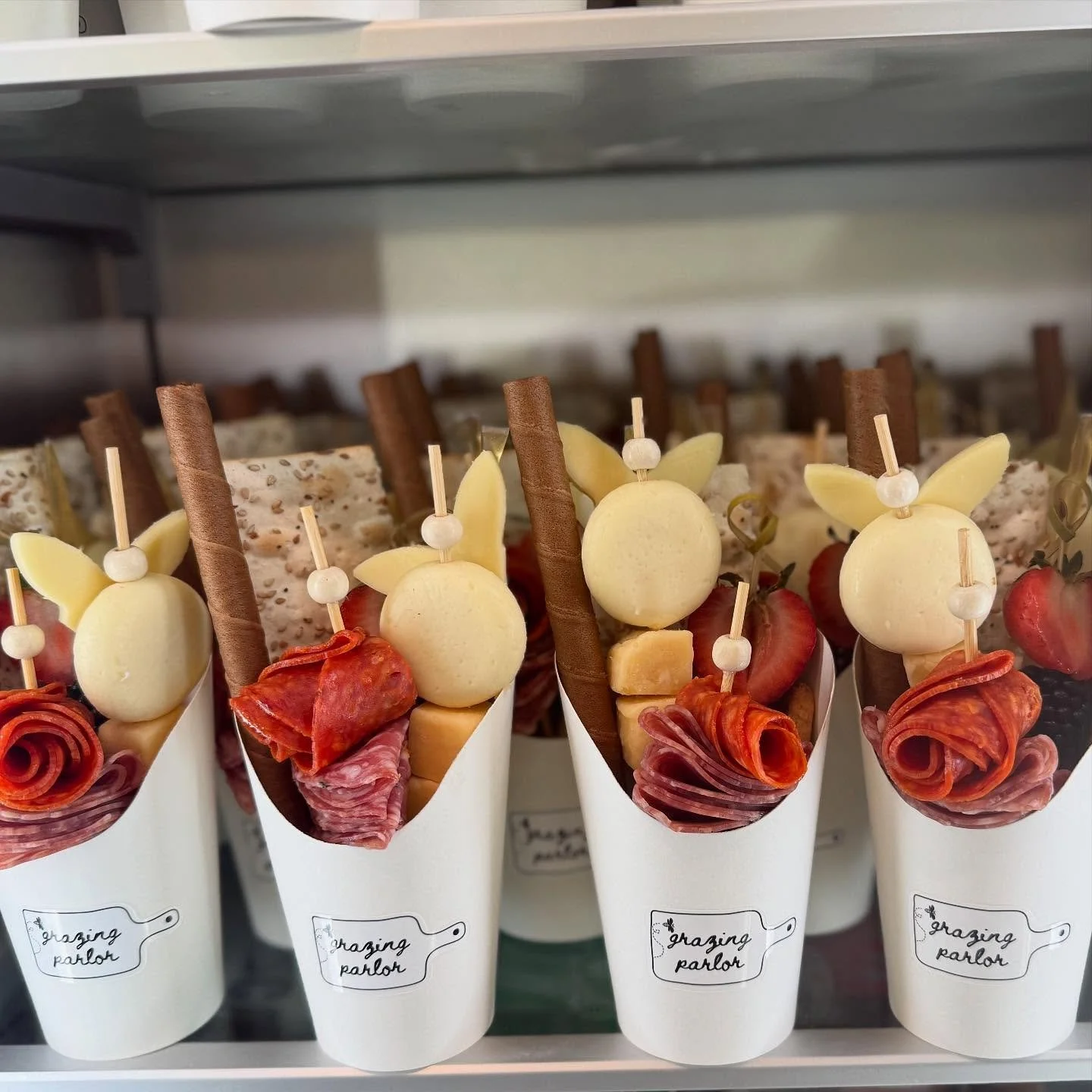 Appetizer or snack cups filled with cheese, cured meats, salami, sliced cheese, breadsticks, and cheese balls, labeled 'Grazing Parlor'.