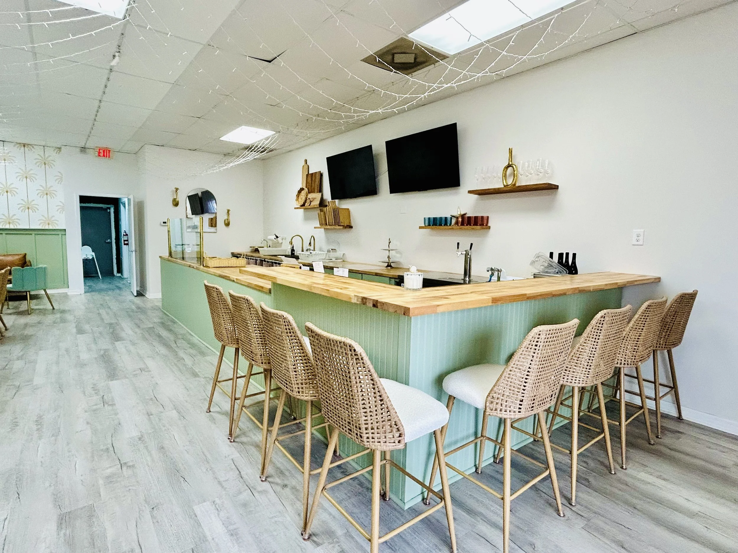 wicker and cream bar stools around a sipping or cheers area where beer, wine, and bubbles are served. Wood charcuterie boards in background, tap, televisions, wine glasses, cups and beautiful upscale tropical decor with gold accents