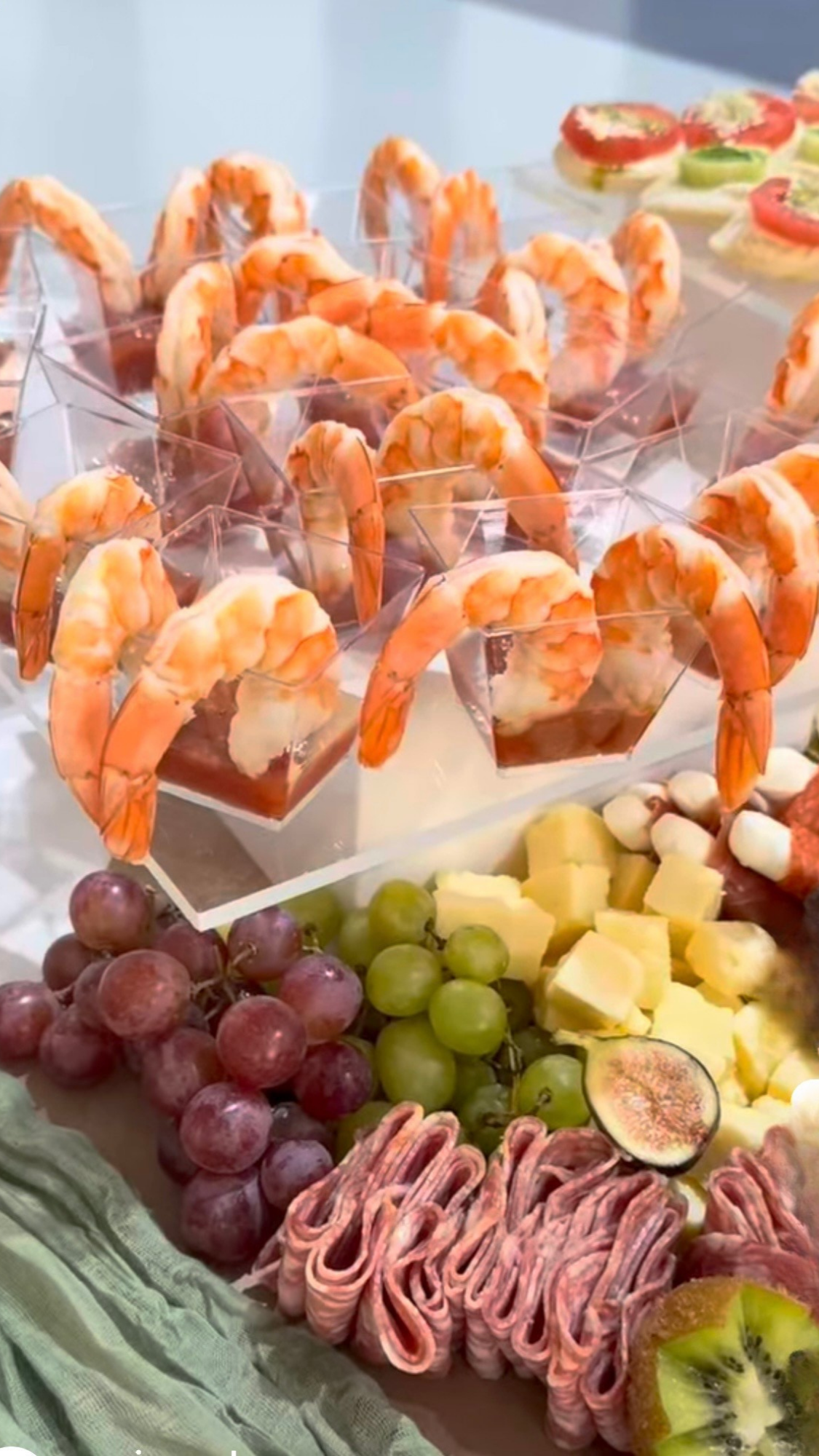 Shrimp cocktail served in small square cups, surrounded by grapes, cheese, sliced fig, ham, and kiwi on a platter.