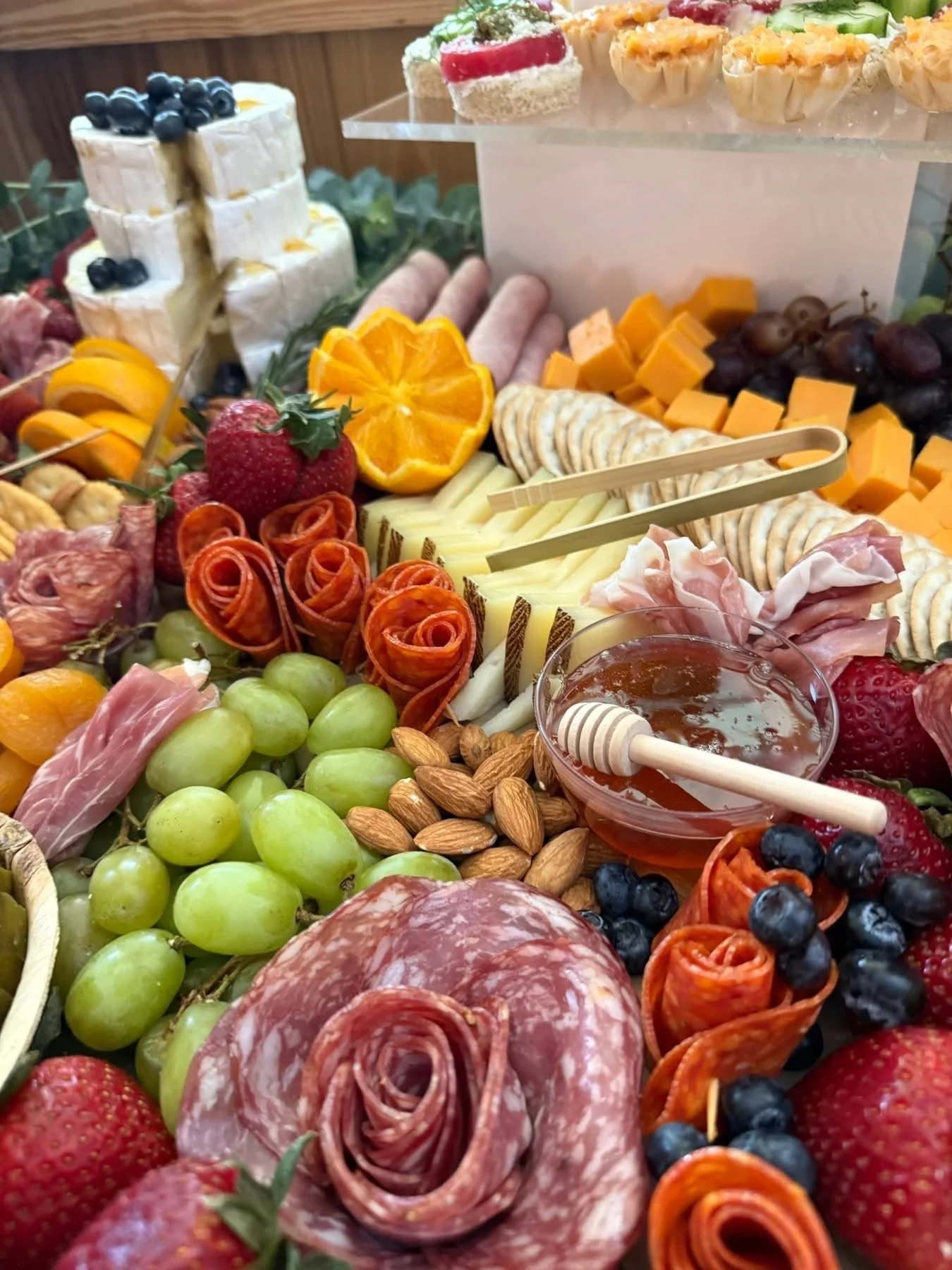 A charcuterie board with cheeses, cured meats, grapes, strawberries, blueberries, almonds, sliced oranges, crackers, and honey, with some sweet treats in the background.