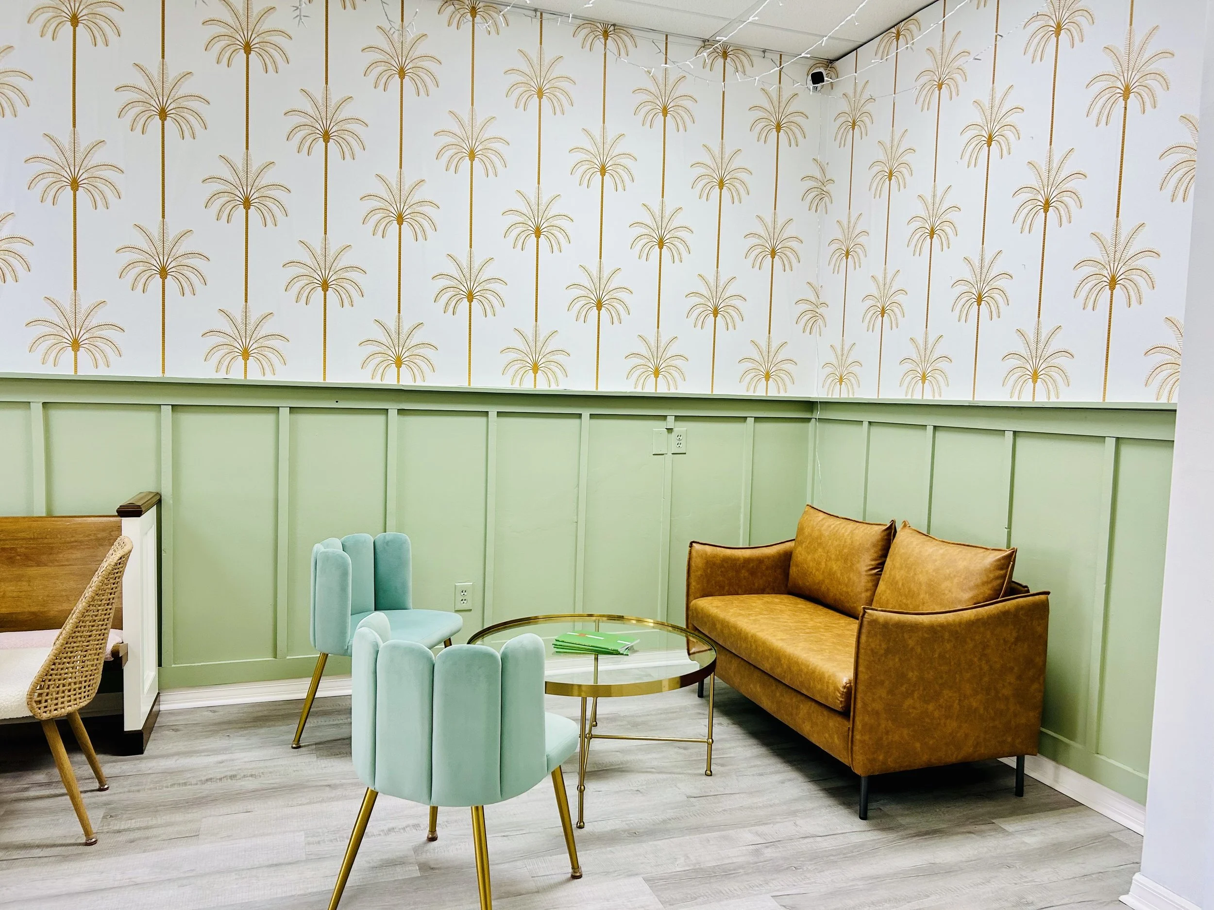 lounge area at Grazing Parlor with comfy leather couch, comfy green aesthetic chairs, glass coffee table with gold accents, mint board and batten wall with tropical wall paper, cream with gold palm trees.