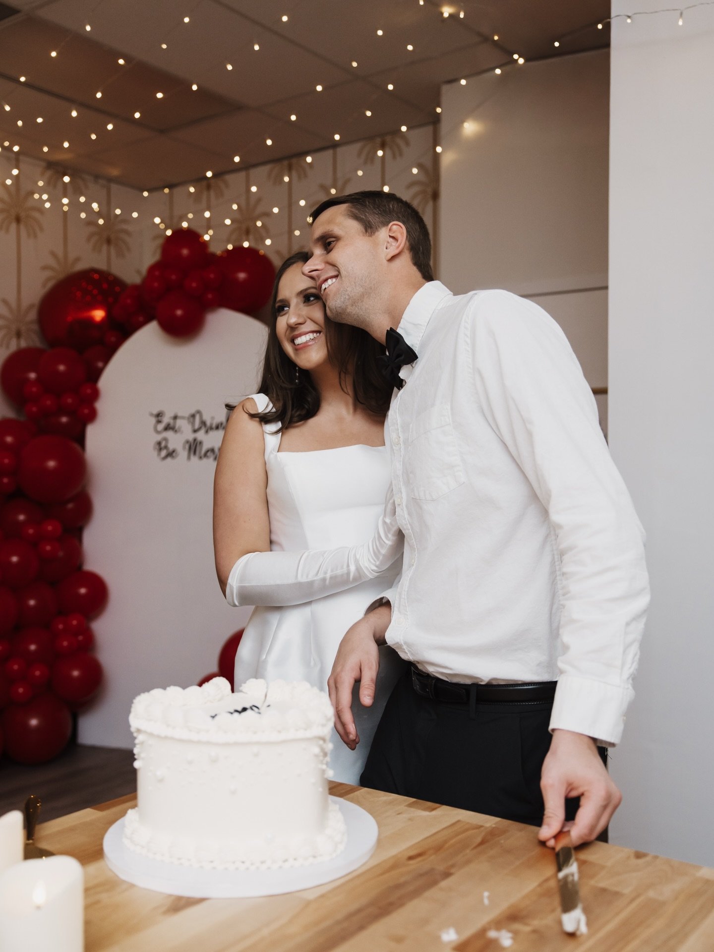 A couple at their wedding reception standing behind a white cake on a wooden table, smiling and posing for photos. The woman is wearing a white wedding dress with gloves, and the man is wearing a white shirt with a black bowtie. The background features decorative lighting, a red balloon arch, and a sign that reads ‘Eat, Drink, Be Married’.