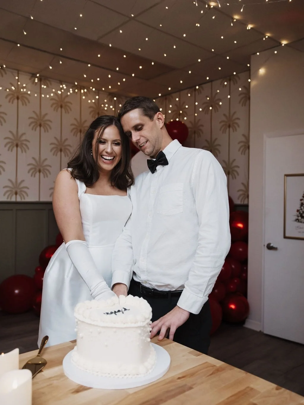 A newlywed couple, smiling and happy, cutting a wedding cake together at their celebration. The bride is wearing a white dress with long gloves, and the groom is dressed in a white shirt with a black bow tie. The background features festive string lights and palm tree wallpaper.