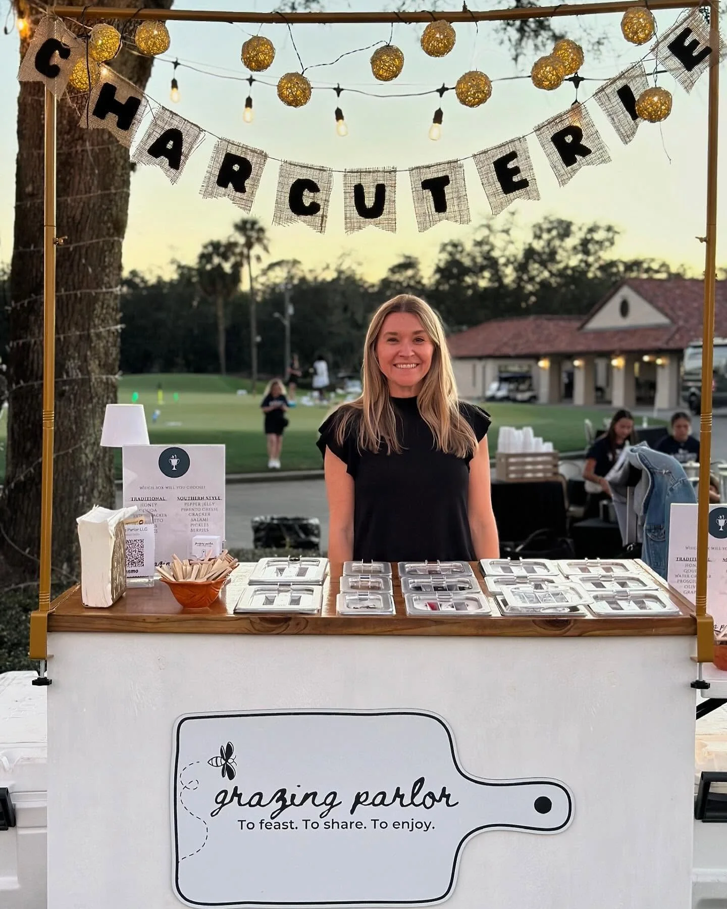 A smiling woman standing behind a table at an outdoor wedding reception, with a sign decorated with fairy lights hanging above. The table features various jewelry displays, and a banner above reads 'CHARTERED E'. The table has a sign that says 'grazing parlor' with a description 'To feast. To share. To enjoy.' The background shows a lawn with people, trees, and a building with a red roof.