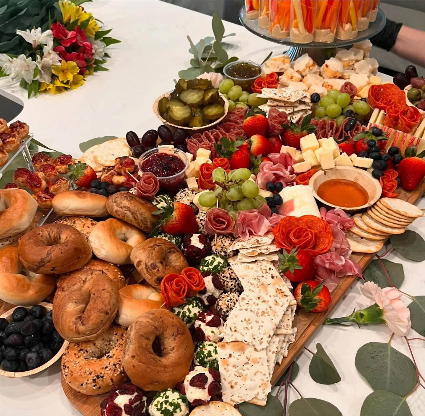 A large charcuterie board with assorted cheeses, meats, fruits, vegetables, pastries, and condiments, surrounded by additional snacks and floral decorations.