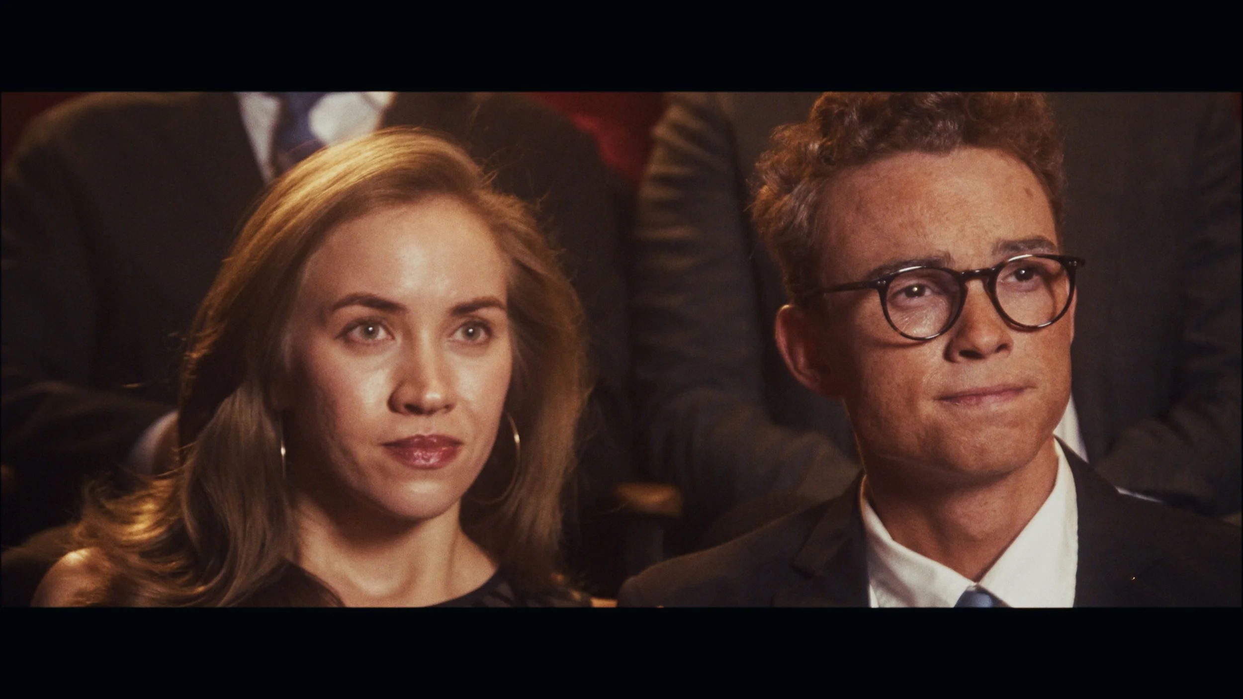 Close-up of a woman with light brown hair and a man with curly hair and glasses, attending a formal event.