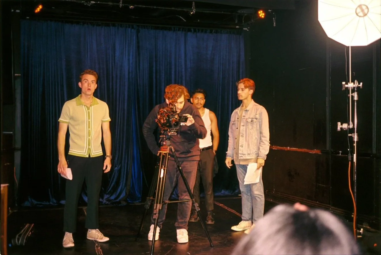 Four men are on a small stage with dark blue curtains. One man is filming or taking a picture with a camera on a tripod. The man on the left is holding papers and appears surprised. The three other men are standing, one holding papers, and all are looking in different directions. There is a large studio light to the right.