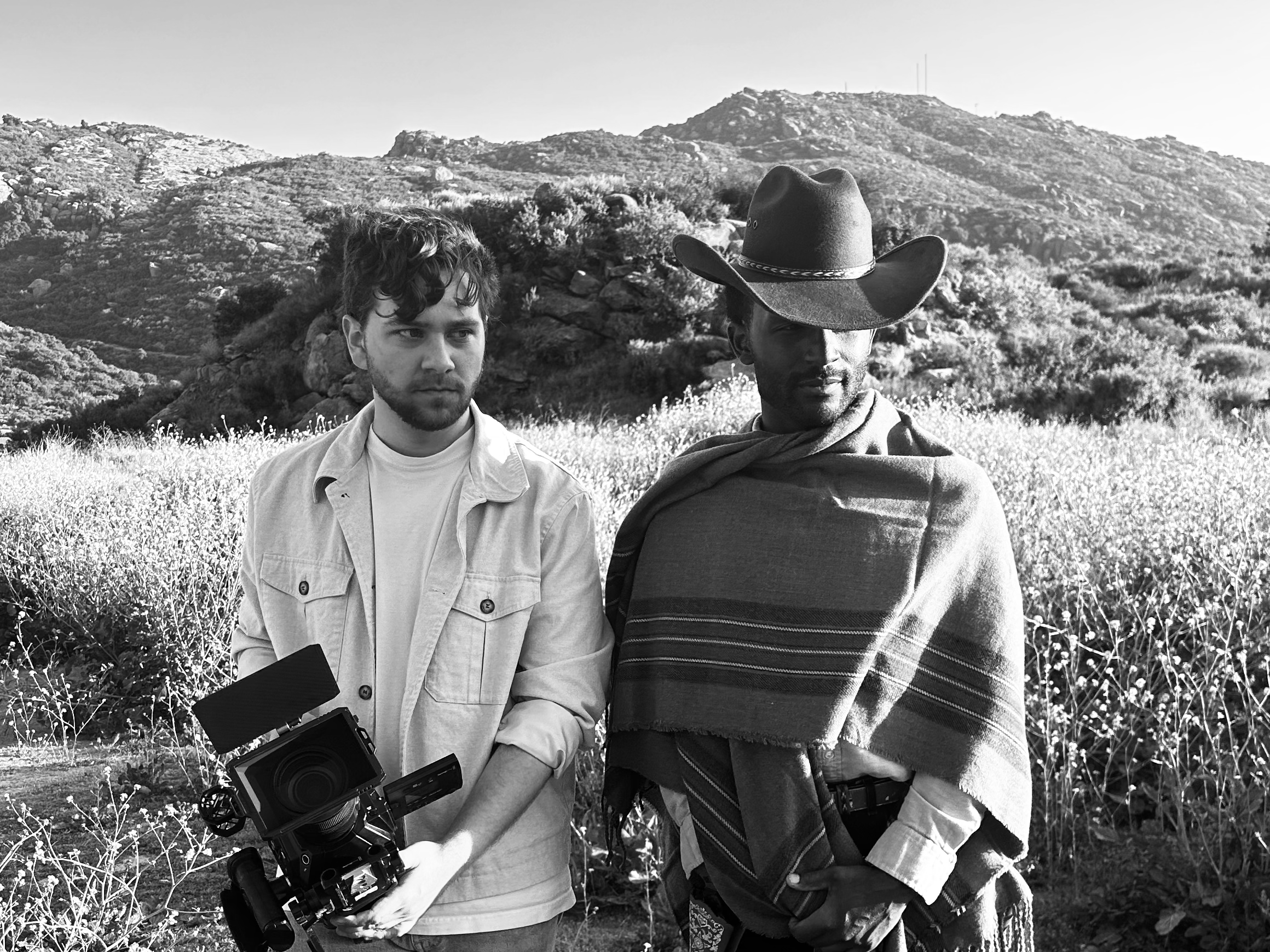Two men outdoors in a hilly landscape with sparse vegetation. One man has curly hair and is holding a camera. The other man is wearing a cowboy hat and a striped shirt.