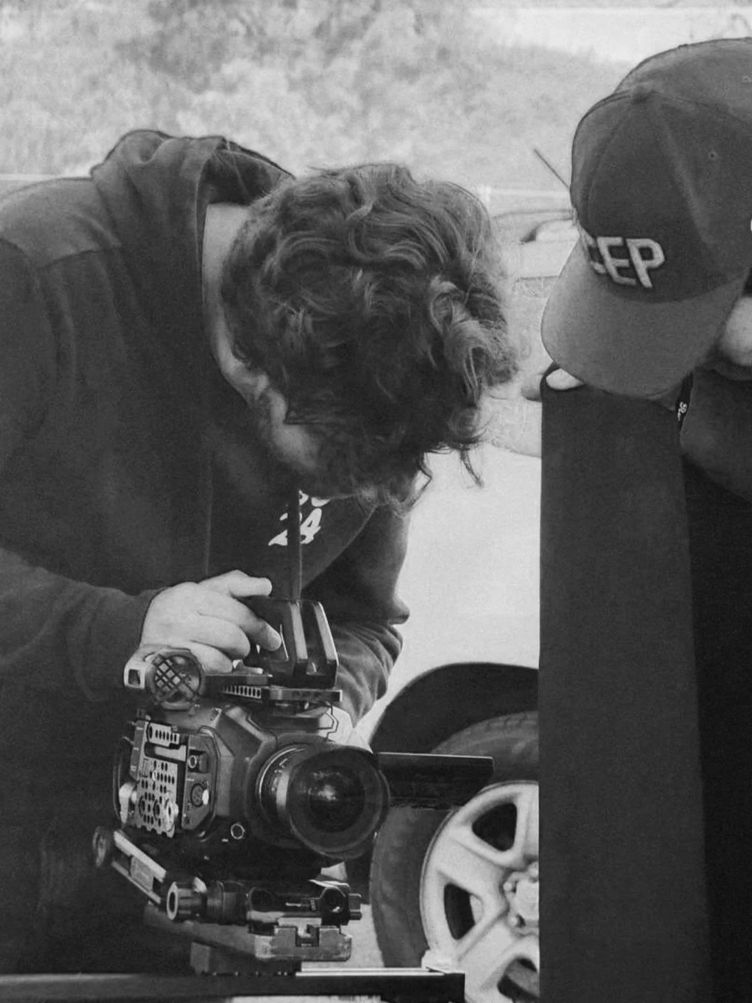 Two filmmakers filming a scene with a professional camera outdoors. One person is wearing a jacket and is looking through the camera, while another is wearing a cap with