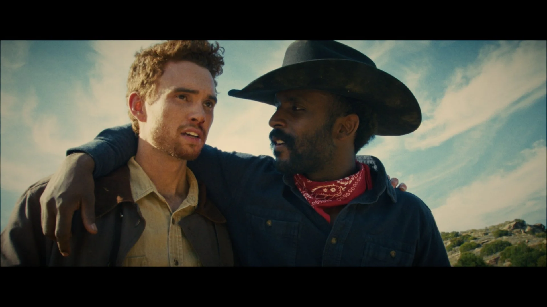 Two men are standing outdoors against a blue sky with some clouds. One man, wearing a cowboy hat and red bandana, has his arm around the other man, who has red hair and freckles. They appear to be talking or sharing a moment.