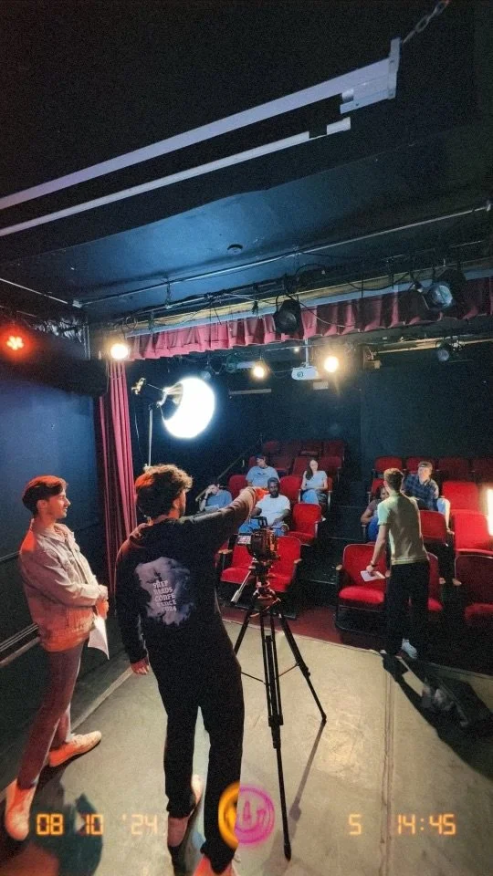 The image depicts a small theater or studio with red seats and black walls. Several people are seated, possibly preparing for a performance or a recording session. One person is adjusting a camera on a tripod, while others are standing or sitting around, some engaging in conversation or preparing. The room is dimly lit with studio lights and a large reflector or softbox directing light towards the scene.