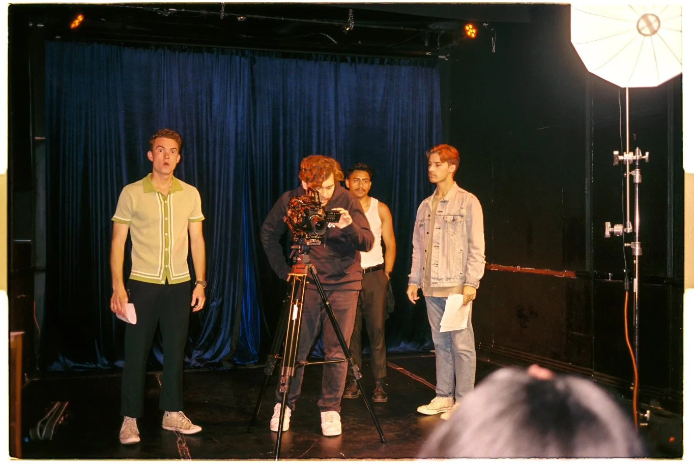 Four young men on a stage with a dark blue curtain backdrop, one man operating a camera and three others standing around, one holding a script, during a filming or rehearsal.
