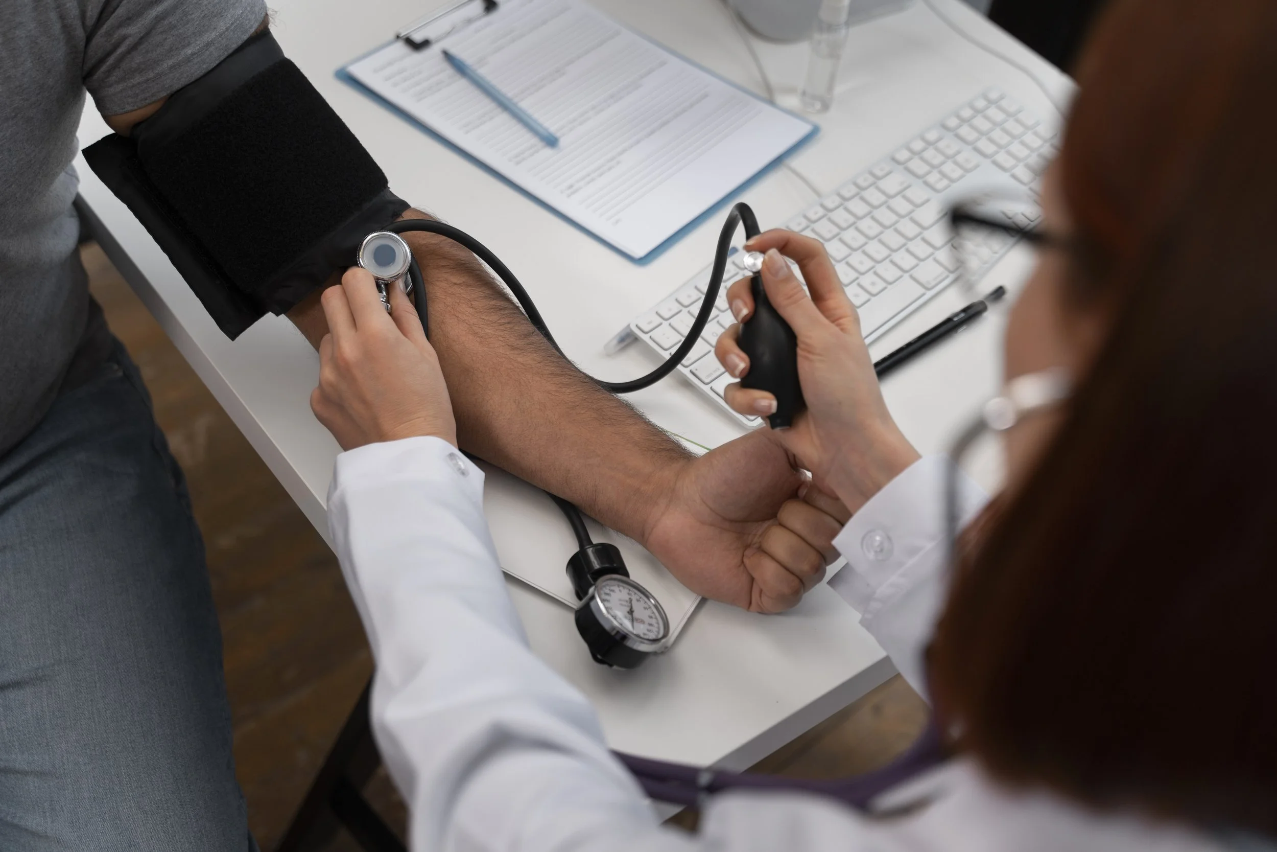 A healthcare professional measuring a patient's blood pressure using a sphygmomanometer and stethoscope, with a computer keyboard, clipboard, pen, and water bottle on the desk.