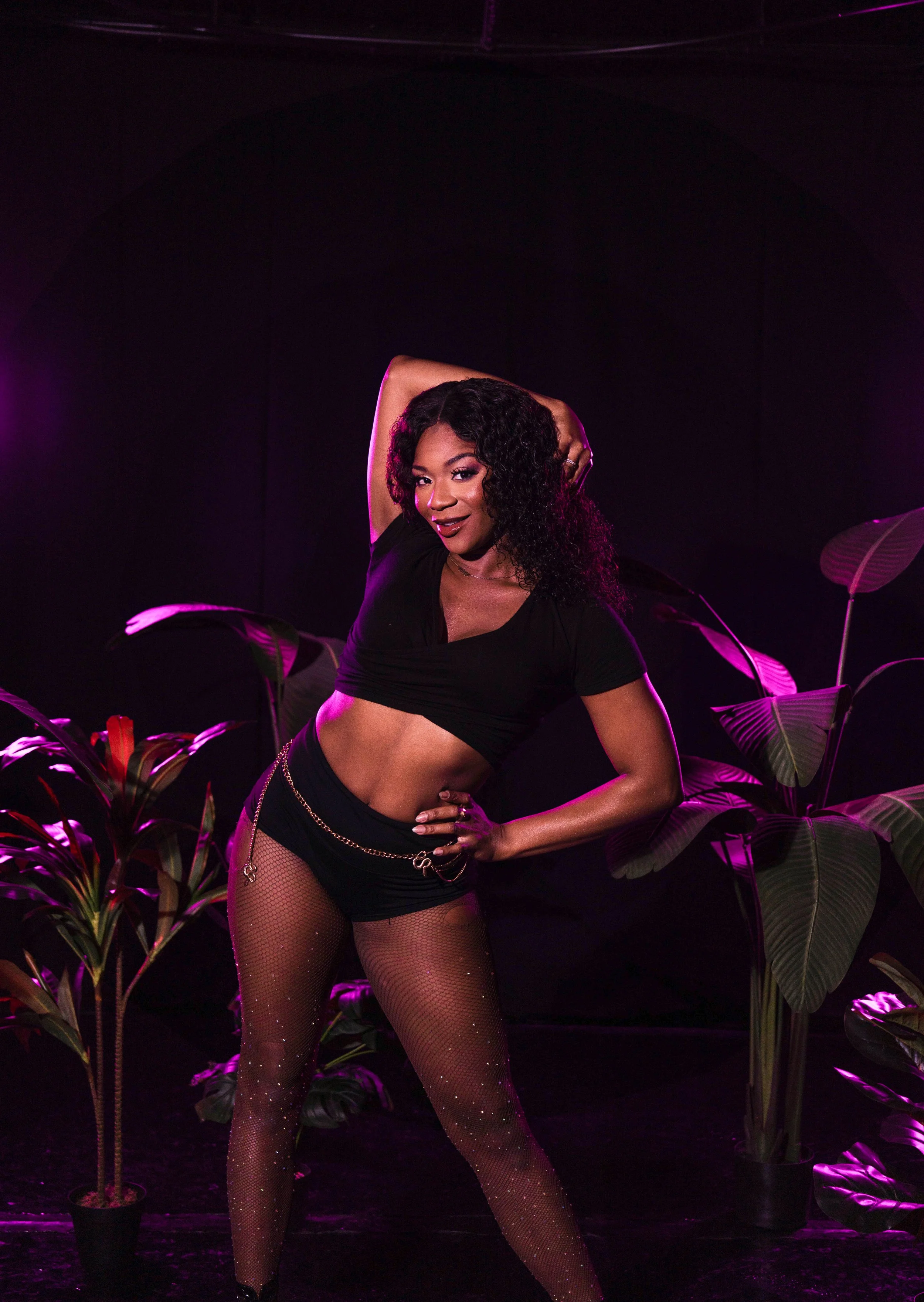 A woman with dark curly hair poses confidently with one arm behind her head and the other on her hip, surrounded by potted plants, in a dark setting illuminated with purple lighting.