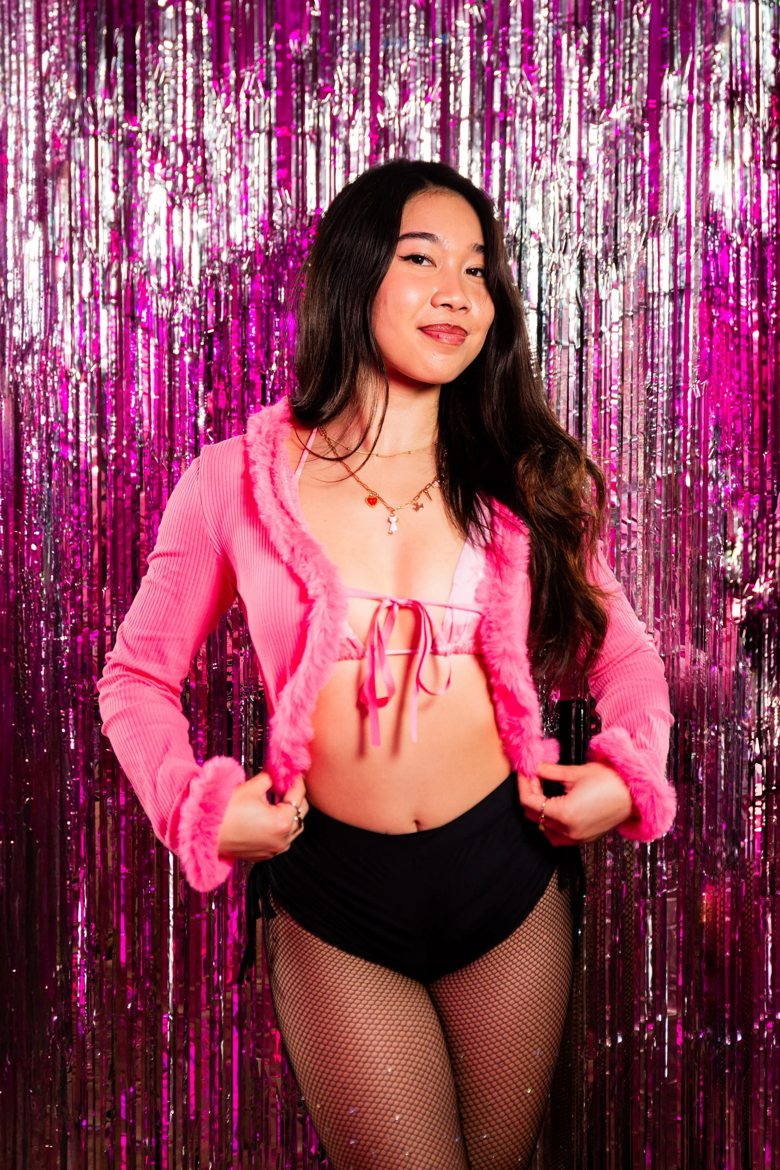 A woman with long dark hair, hoop earrings, and a nose ring poses with one hand behind her head, wearing a pink feathery top, and a black skirt, with pink and purple lighting.