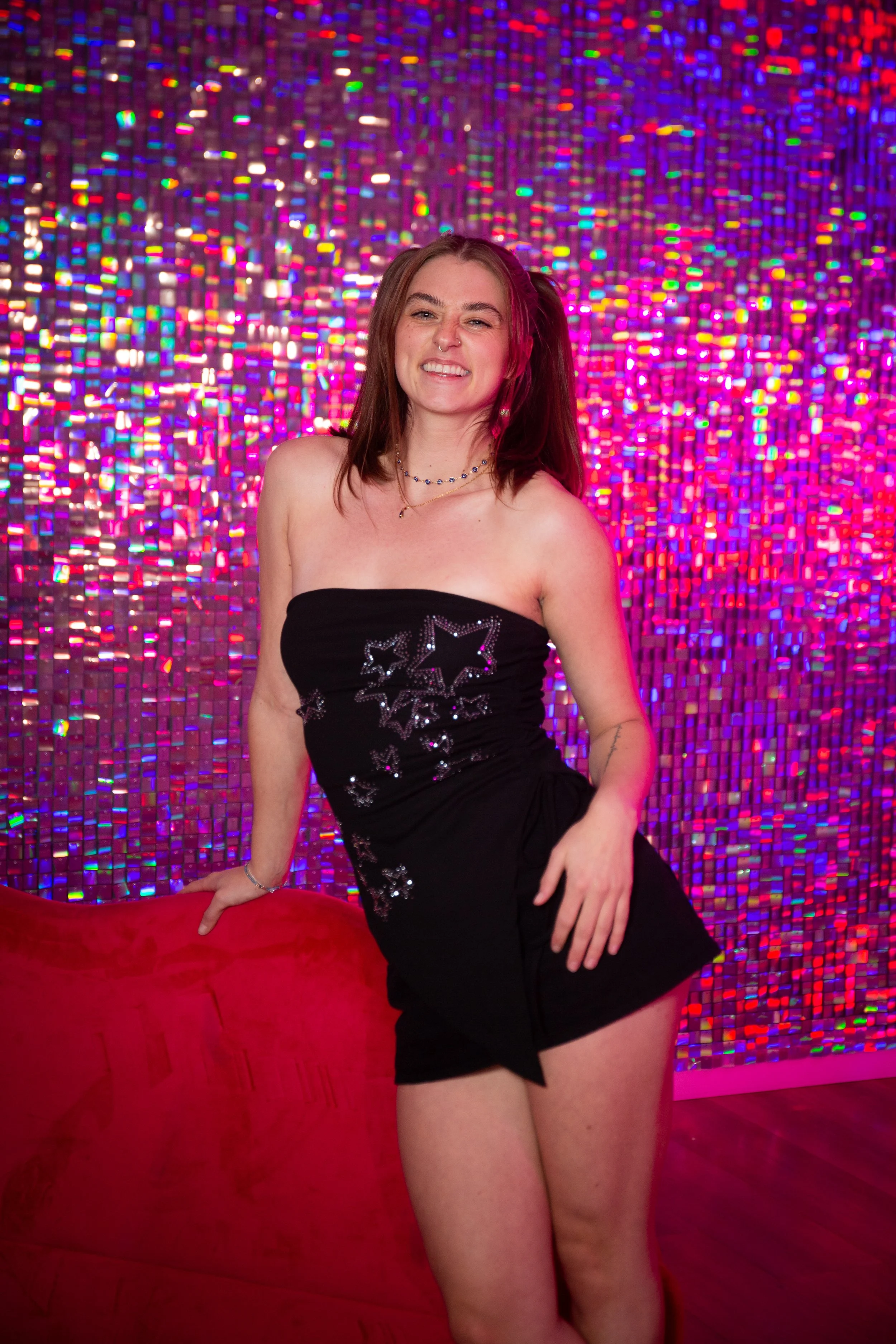 a young woman in a y2k outfit that is a black mini skirt and tube top with sunglasses. Posing against a red lips couch and iridescent shimmery backdrop