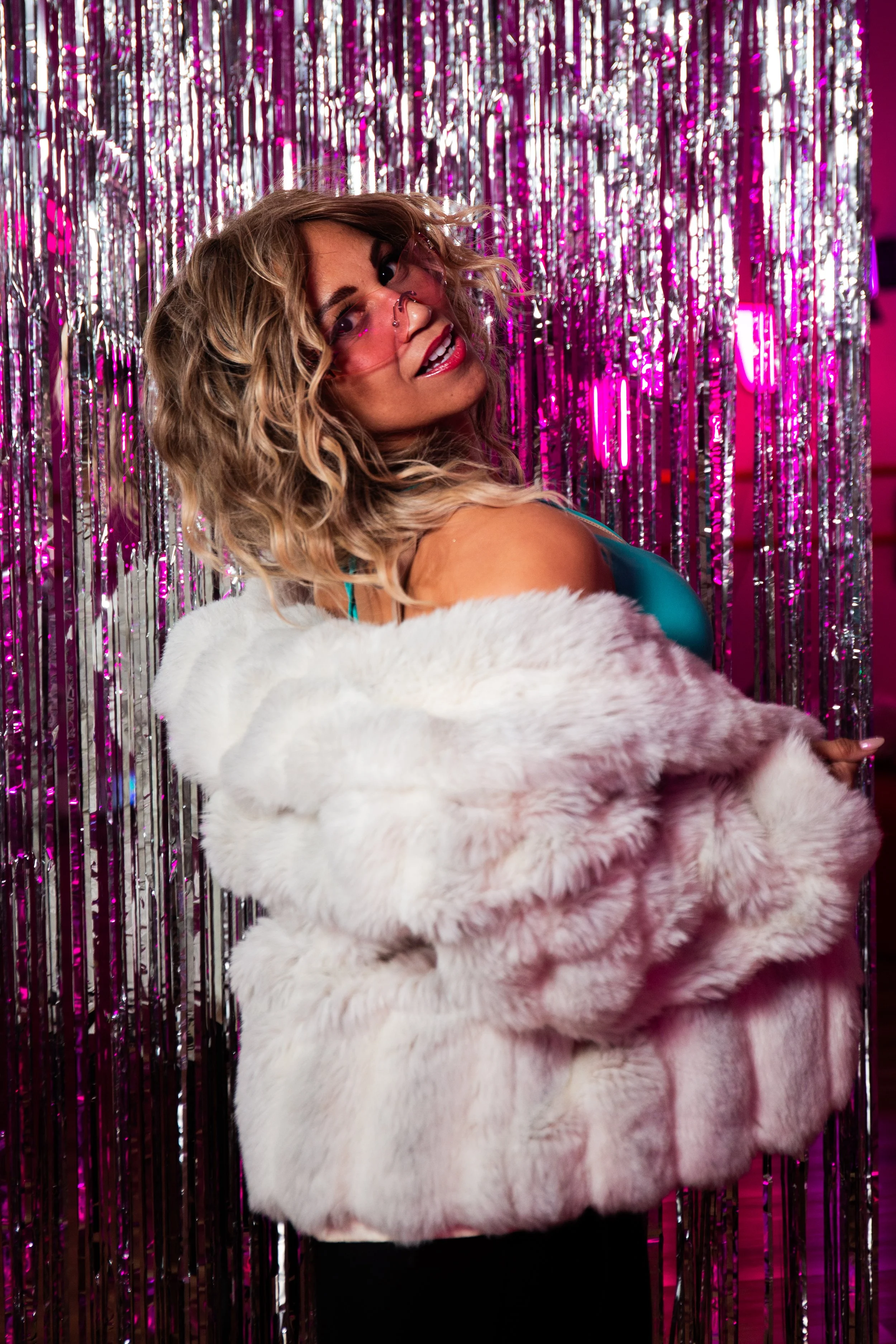 a woman in a white fur coat and turquoise bra. Blonde wavy hair, sunglasses next to a shimmery backdrop