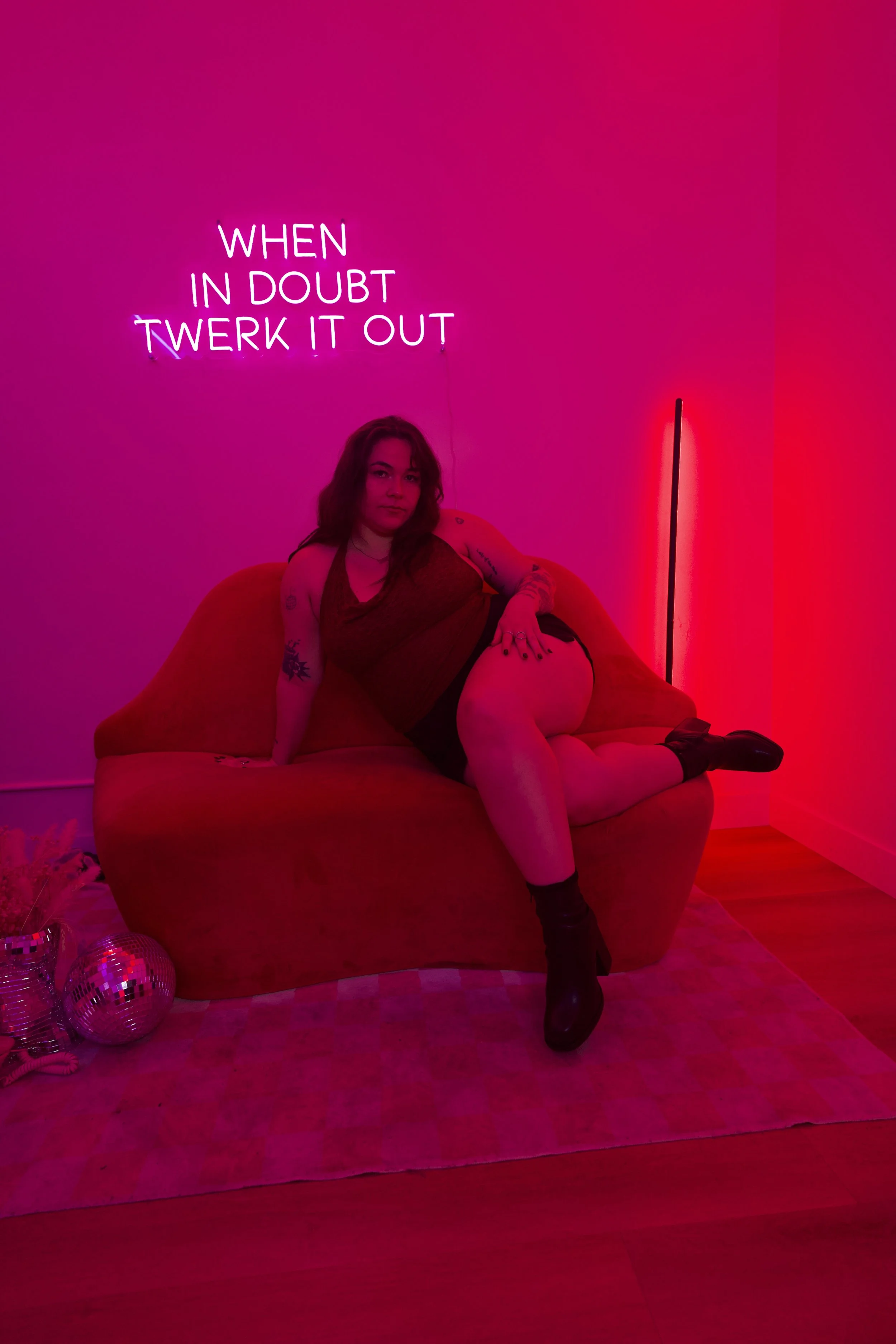 A woman with long dark hair, wearing a green jacket, black shorts, and black high-heeled shoes, crouching and looking at the camera against a plain wall.