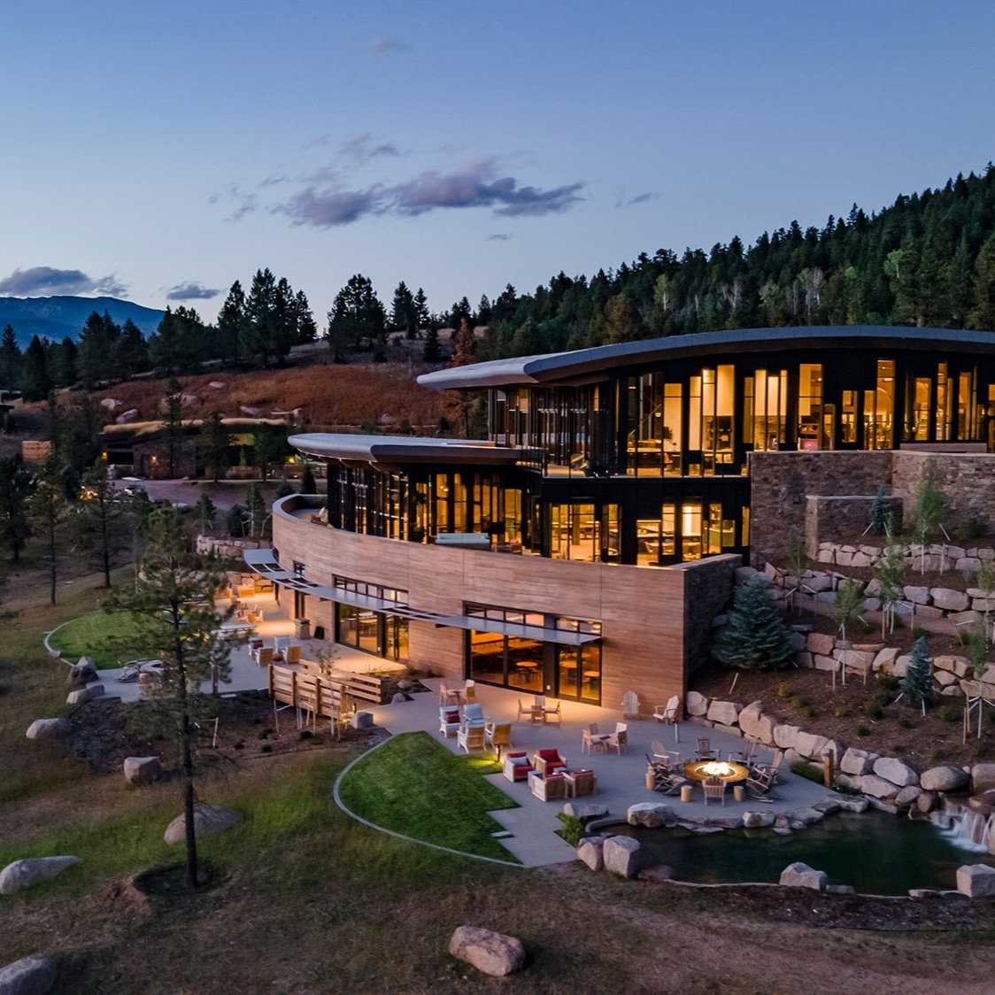 Modern hillside building with large windows, surrounded by forest, patio with seating and fire pit in foreground.