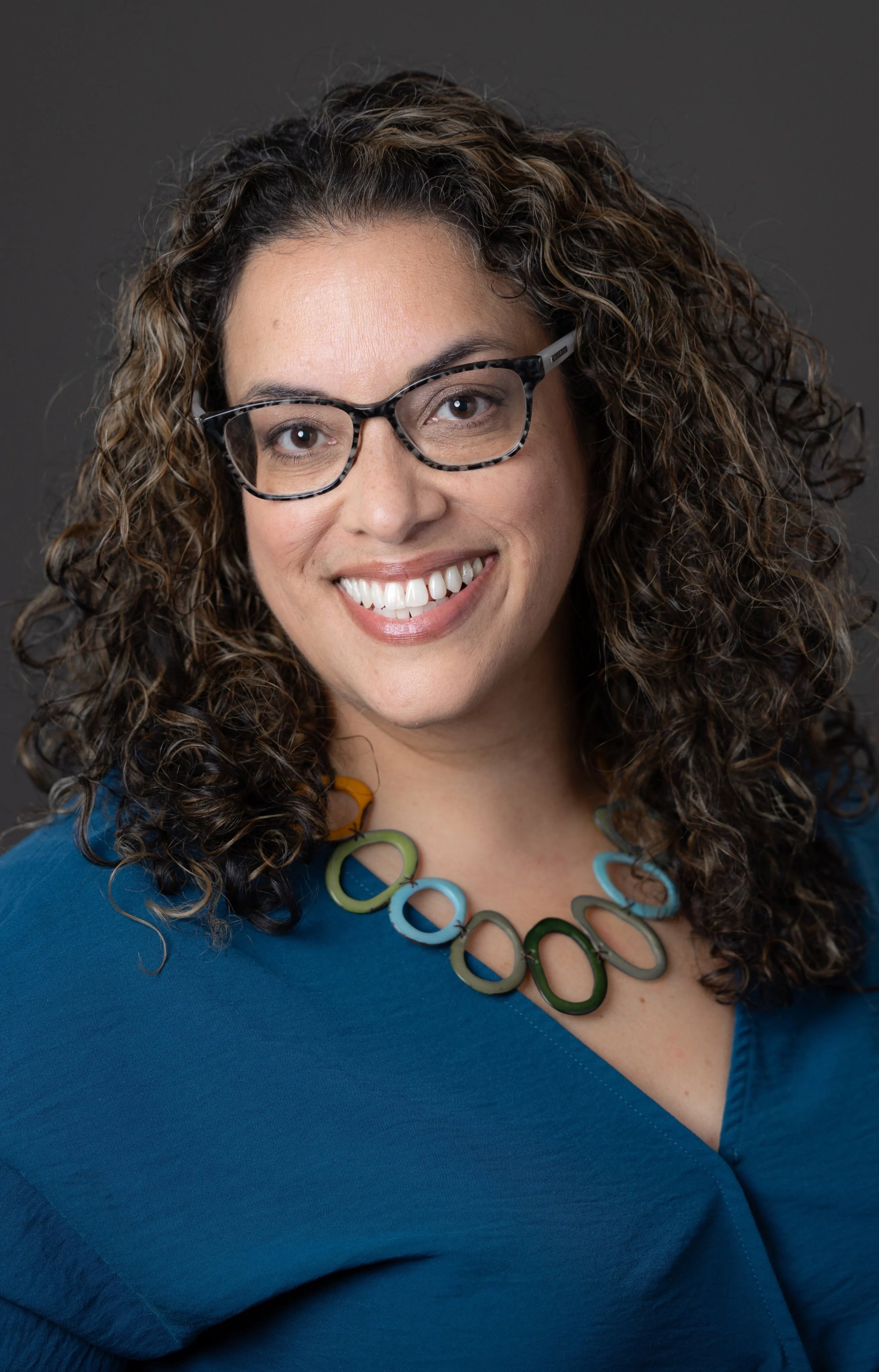 A smiling person with curly hair, wearing glasses and a blue top, accessorized with a colorful necklace.