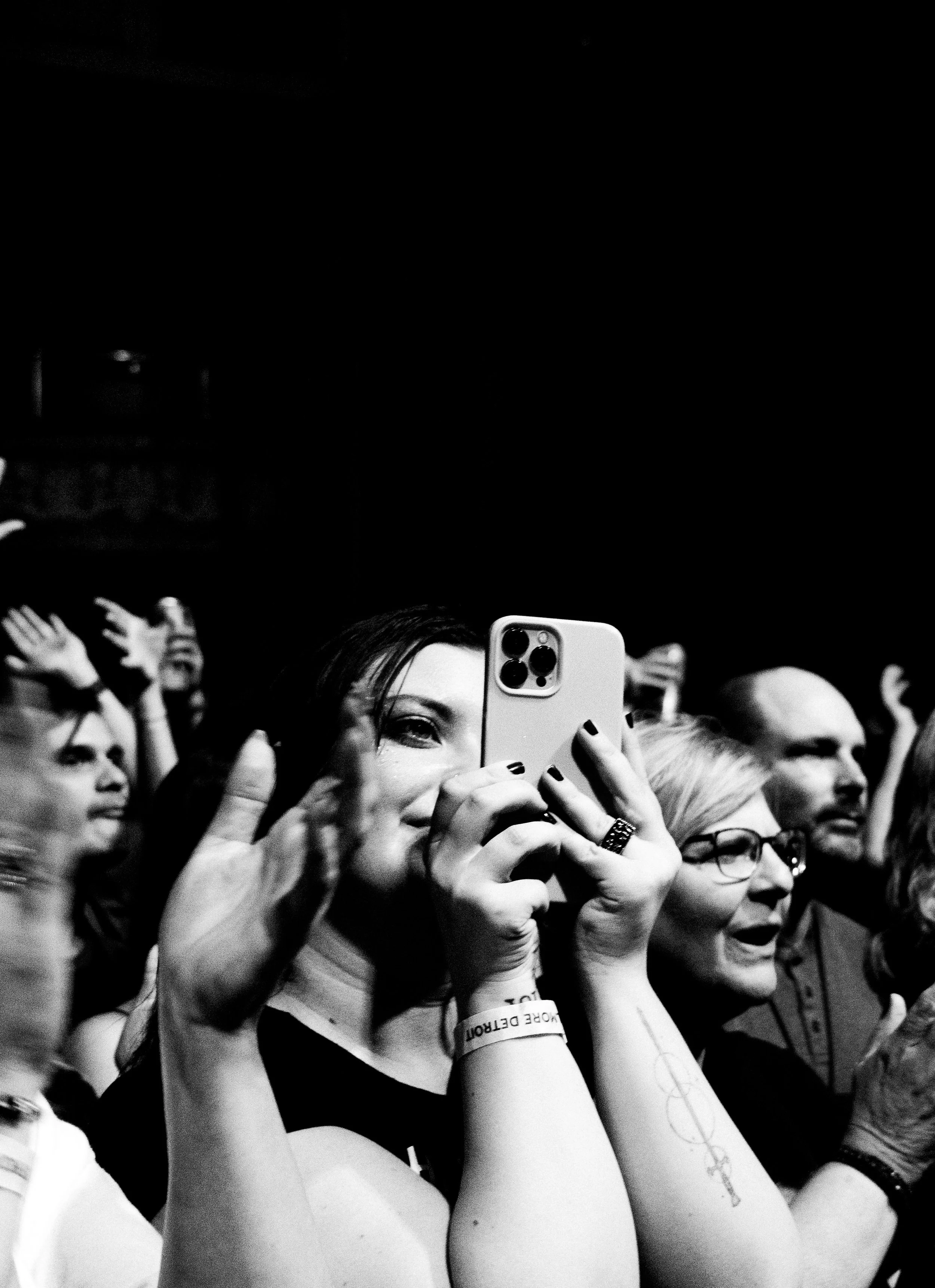 A group of people at a concert or event, with a woman in the foreground taking a selfie or photo with her phone, smiling and wearing a wristband.