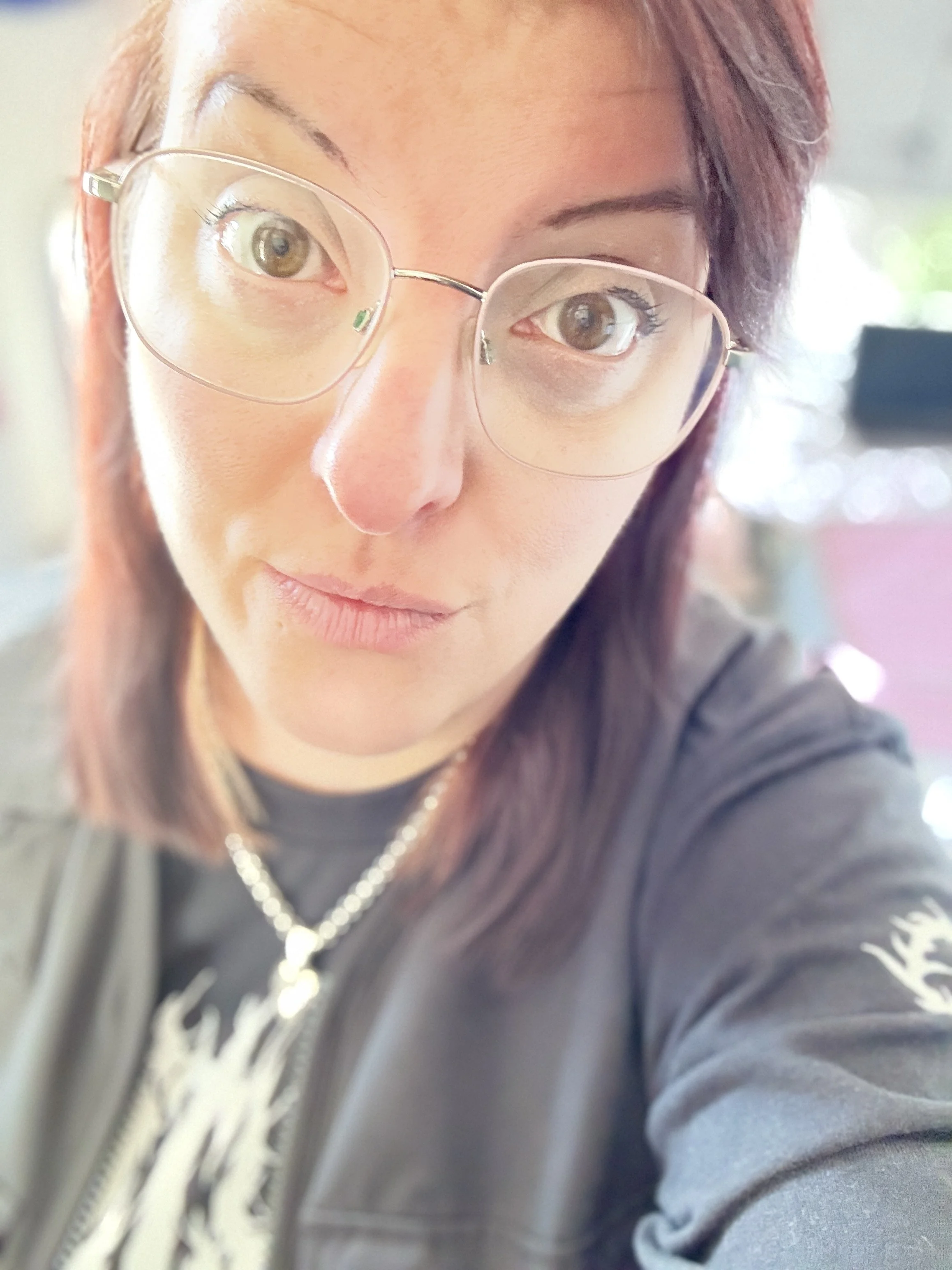 A woman with reddish-brown hair wearing glasses, a necklace, and a black top, taking a close-up selfie.