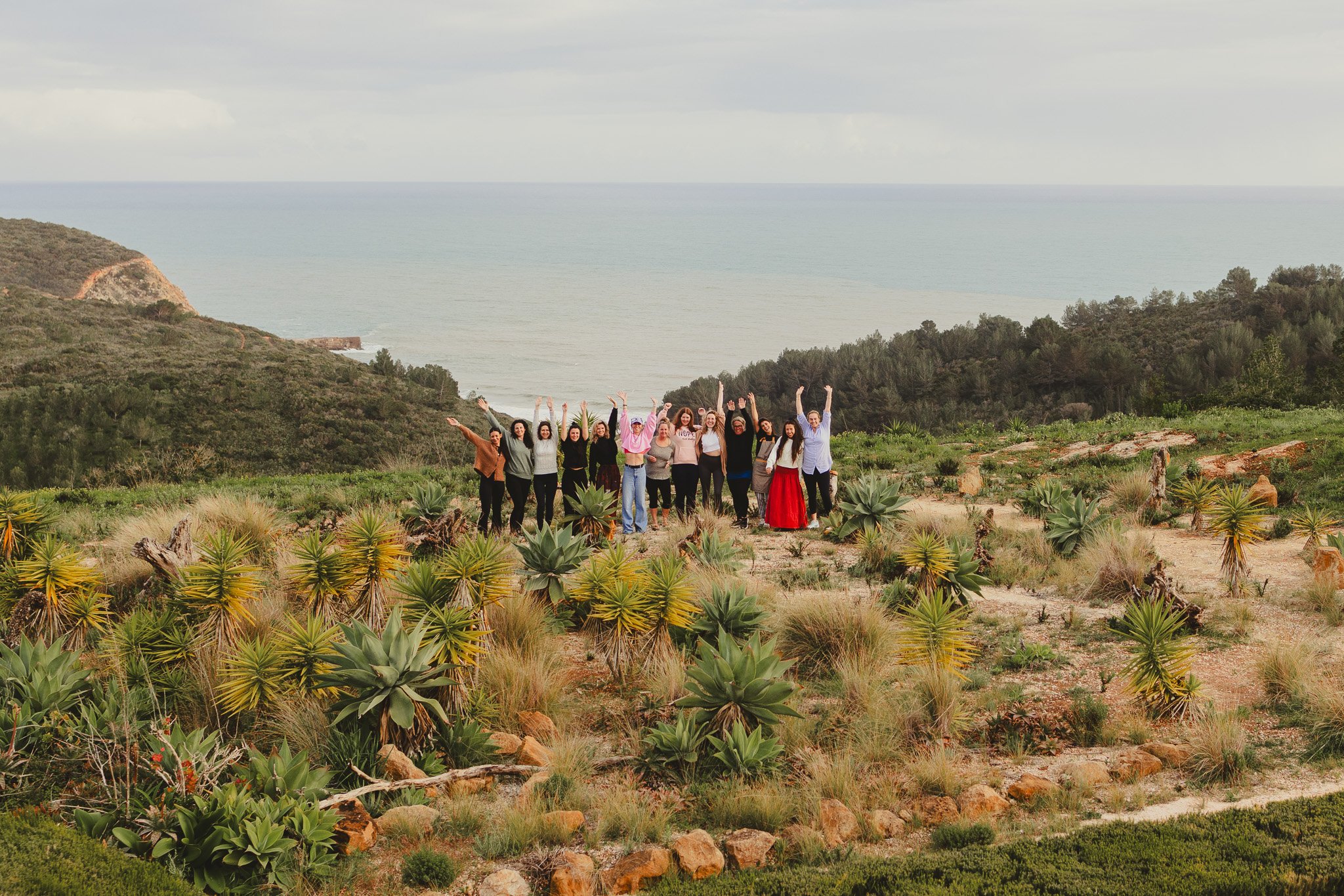 A group of people standing in a desert landscape with drought-tolerant plants, hills, and a body of water in the background, raising their hands and smiling.