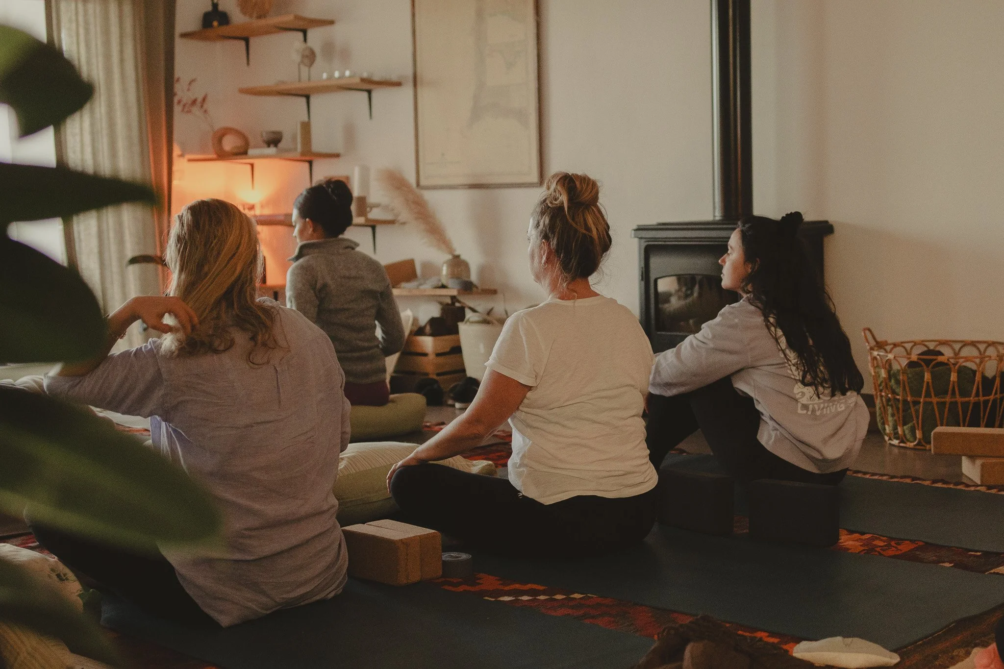 Four women sitting cross-legged on floor cushions in a cozy living room, engaging in a meditative or mindfulness practice. The room has warm lighting, a stove, shelves with decor, and a framed map on the wall.