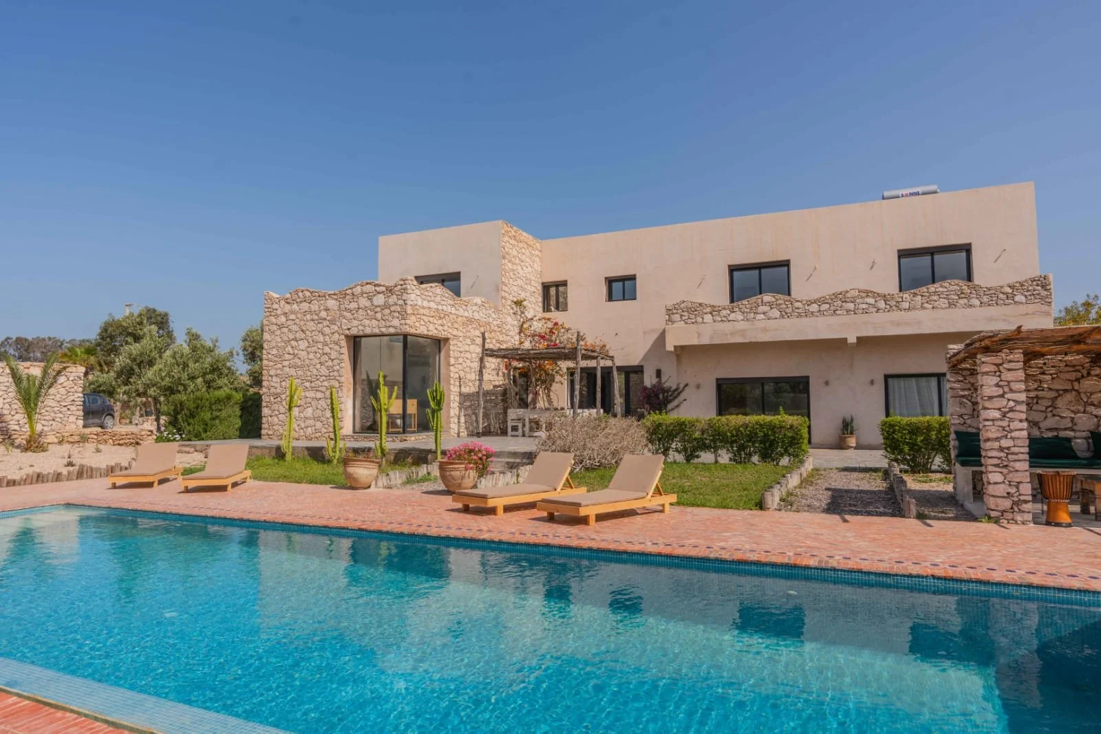 Modern house with stone and stucco exterior, large windows, a swimming pool with lounge chairs, potted plants, and a landscaped yard under a clear blue sky.