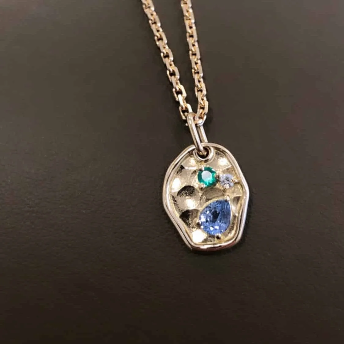 Collier avec pendentif en or 18 carats, incrusté de trois pierres précieuses de différentes tailles et couleurs : bleu, vert et clair, sur fond noir.