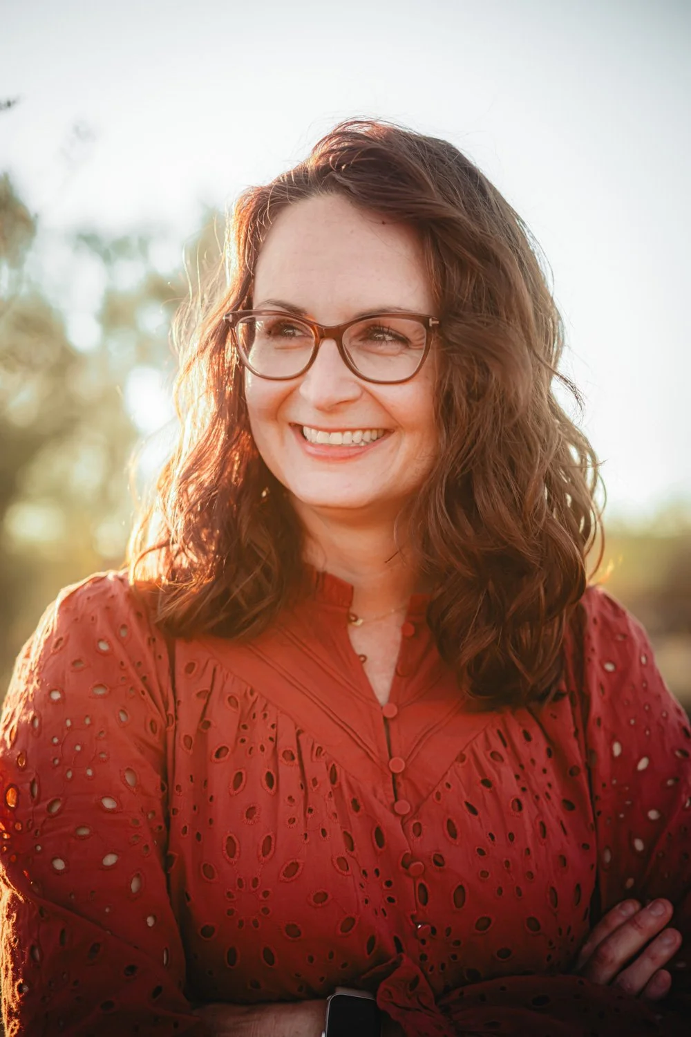 Woman with glasses smiling, wearing a red eyelet blouse, outdoors during sunset.