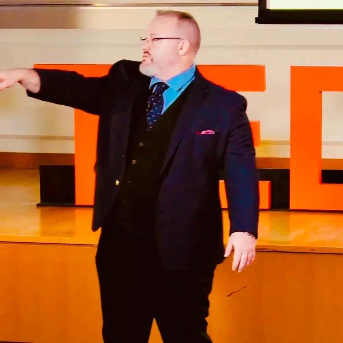 Gentry delivering his TEDx talk, pointing into the audience in front of the orange TED letters.