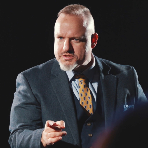 Gentry in a three-piece suit, mid-sentence and gesturing, performing for a corporate audience in a darkened room.