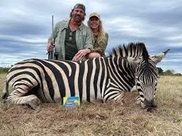 A man and woman posing with a zebra on the ground in a field, under a cloudy sky.