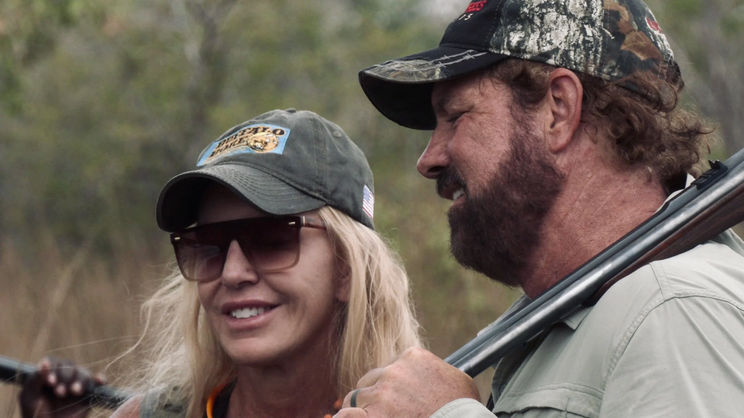 Two people wearing caps outdoors, one holding a rifle over their shoulder.
