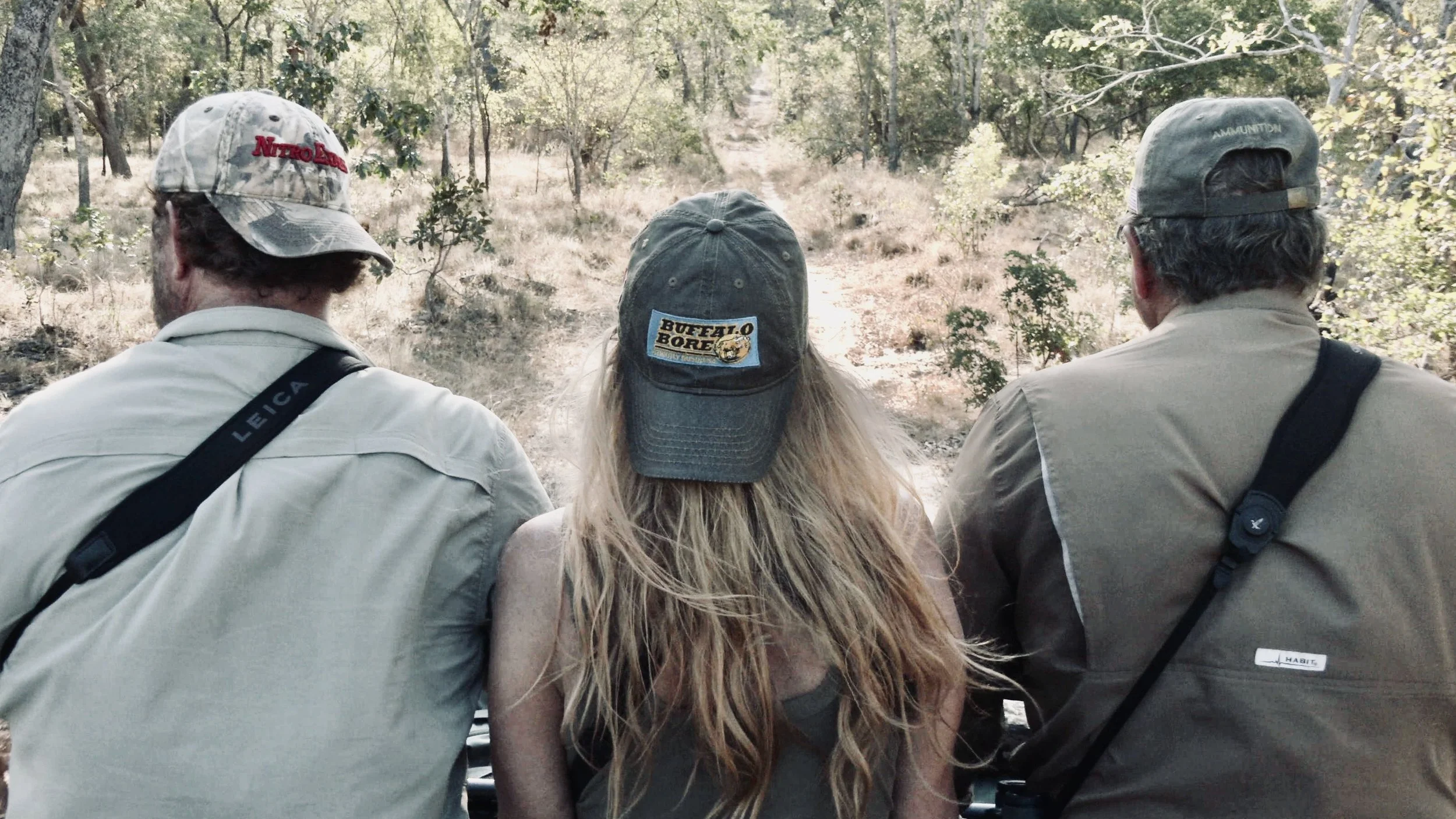 Three people wearing caps, viewed from behind, in a forest setting. The person in the middle has long hair, and the two on the sides are wearing cameras or binoculars over their shoulders.