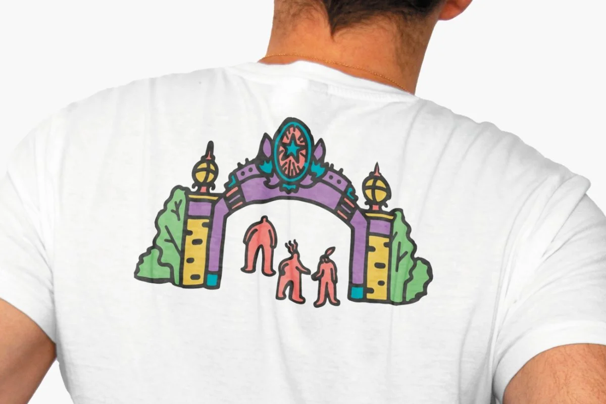 A man wearing a white t-shirt with a colorful drawing of three figures inside an ornate archway with trees on either side.