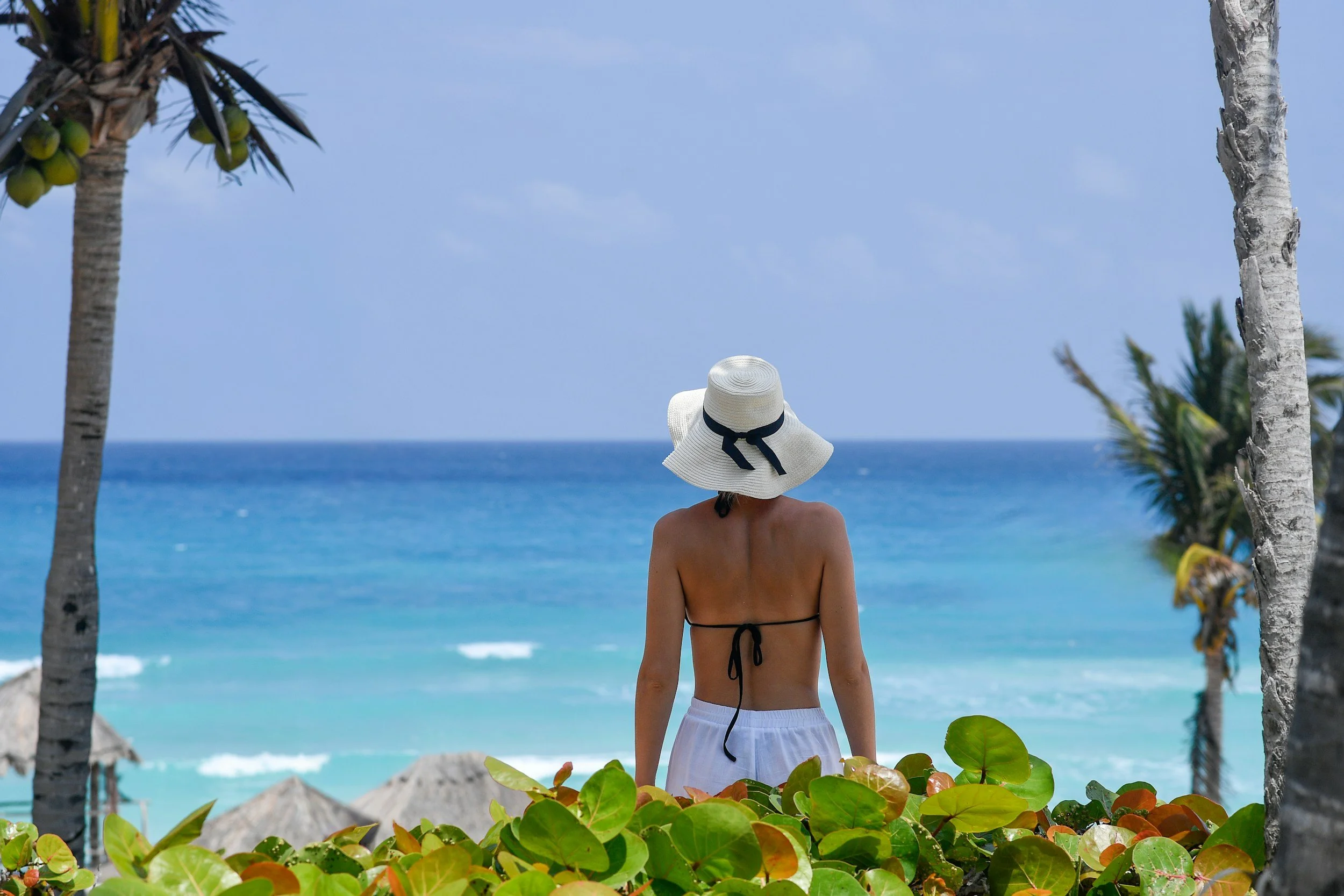 A woman with a wide-brimmed hat looking at the ocean on a tropical beach with palm trees and thatched umbrellas.