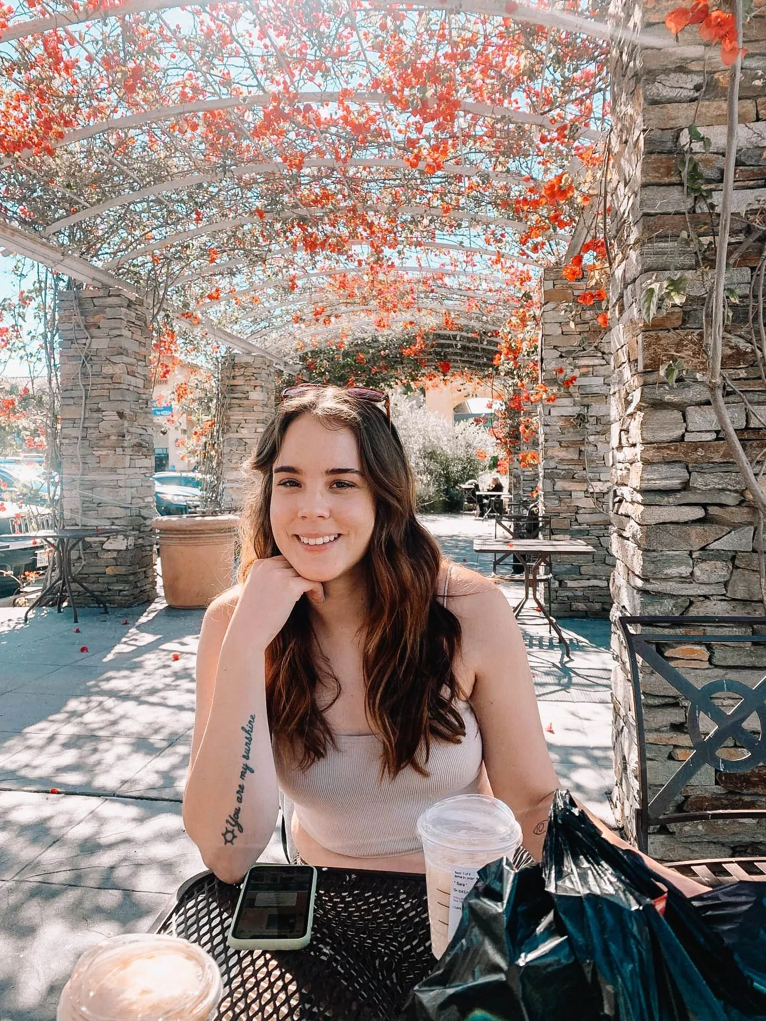 A young woman with brown hair sitting outdoors at a table under a decorative archway covered with orange and red flowers. She is smiling, wearing a light-colored tank top, and holding a beverage cup. On the table are two large drinks, a smartphone, and snack bags. Background includes trees, chairs, and parts of a building.